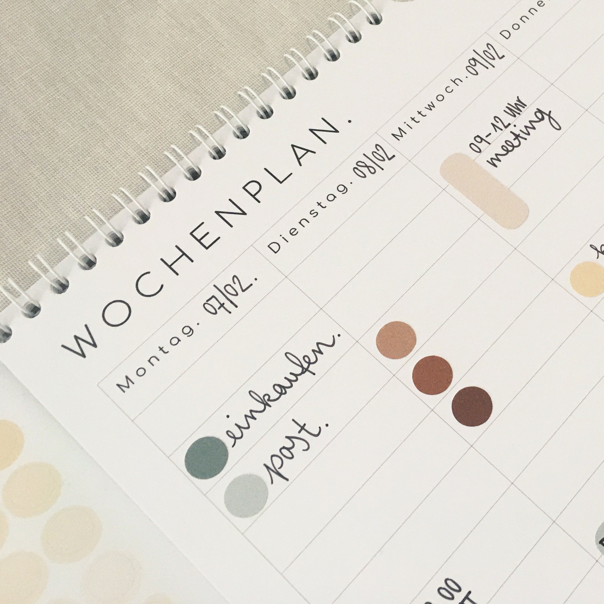 Ein spiralgebundener Wochenplaner in deutscher Sprache zeigt handgeschriebene Pläne für Montag und Dienstag, darunter Einkäufe, Post und ein Uni-Treffen, wobei farbige Punkte die Aktivitäten markieren.