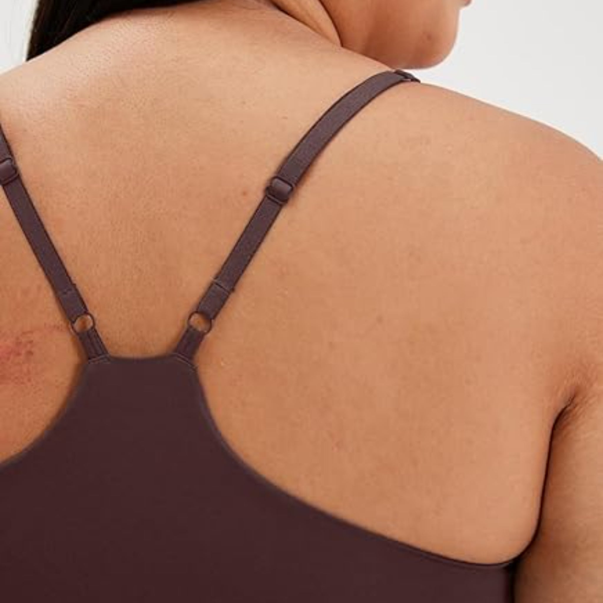 Nahaufnahme des oberen Rückens und der Schultern einer Person, die ein lilafarbenes Tanktop oder einen BH trägt, mit verstellbaren Trägern und einer sichtbaren Narbe oder einem Fleck auf dem linken Schulterblatt.