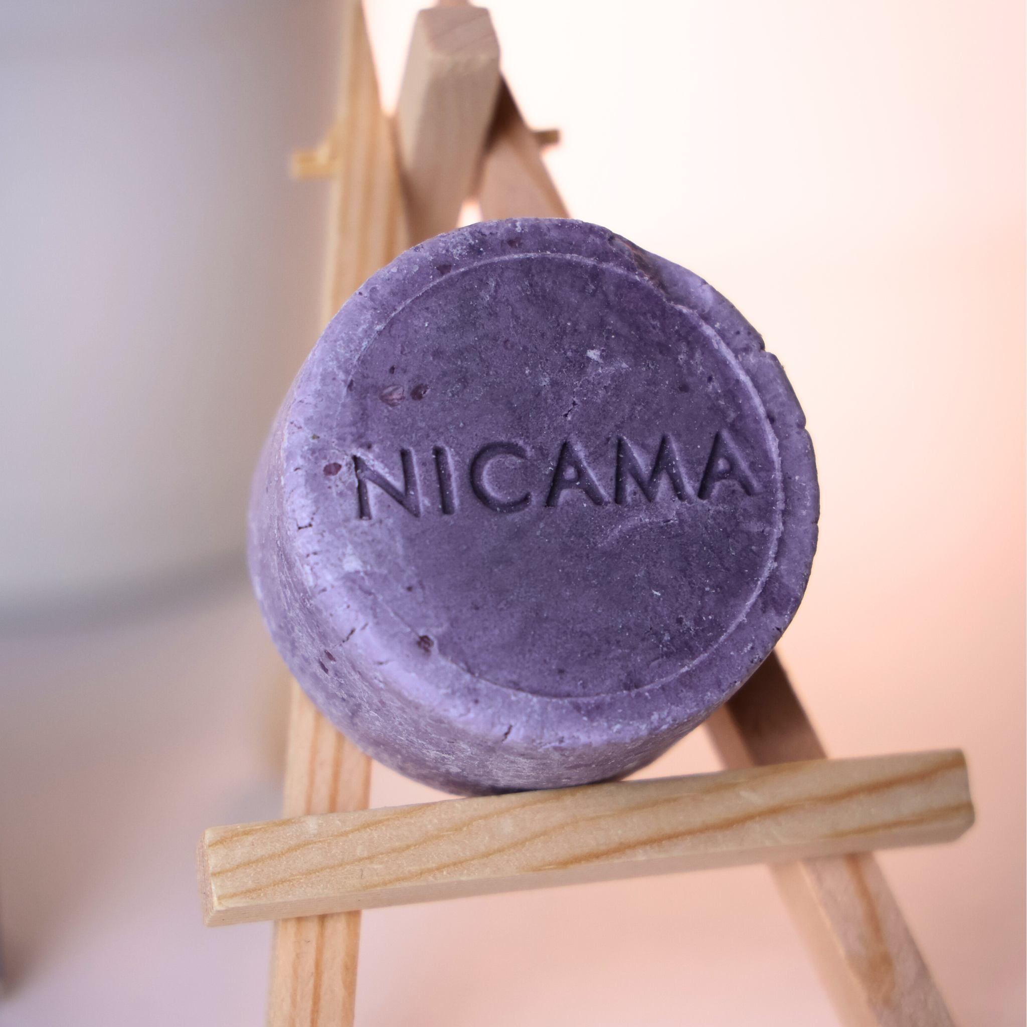 Lila Seifenstück mit der Prägung „NICAMA“ auf einem kleinen Holzständer vor unscharfem Hintergrund.