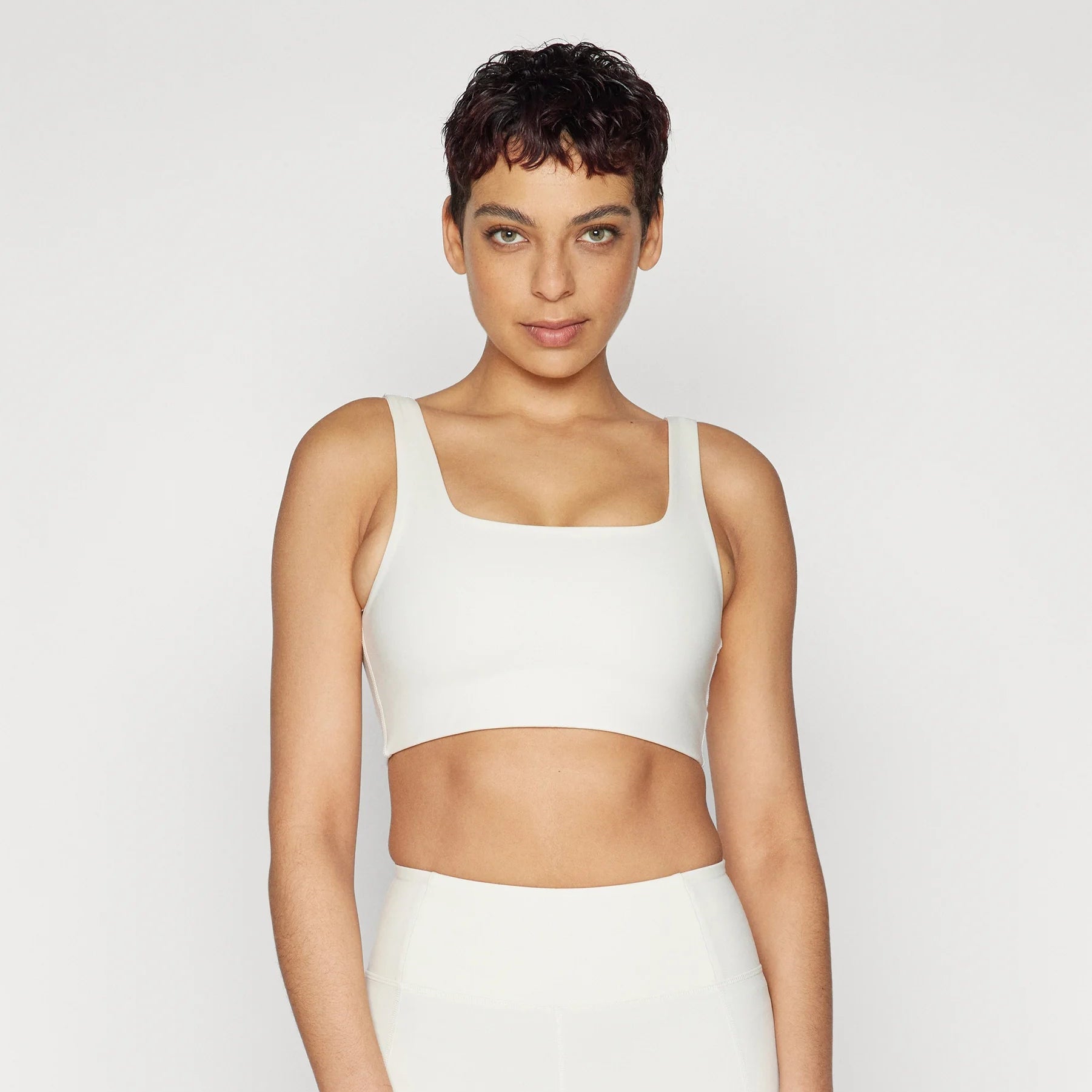 Eine Person mit kurzen dunklen Haaren, die einen weißen Sport-BH und hochgeschlossene weiße Sporthosen trägt, steht vor einem schlichten hellgrauen Hintergrund und blickt selbstbewusst in die Kamera.