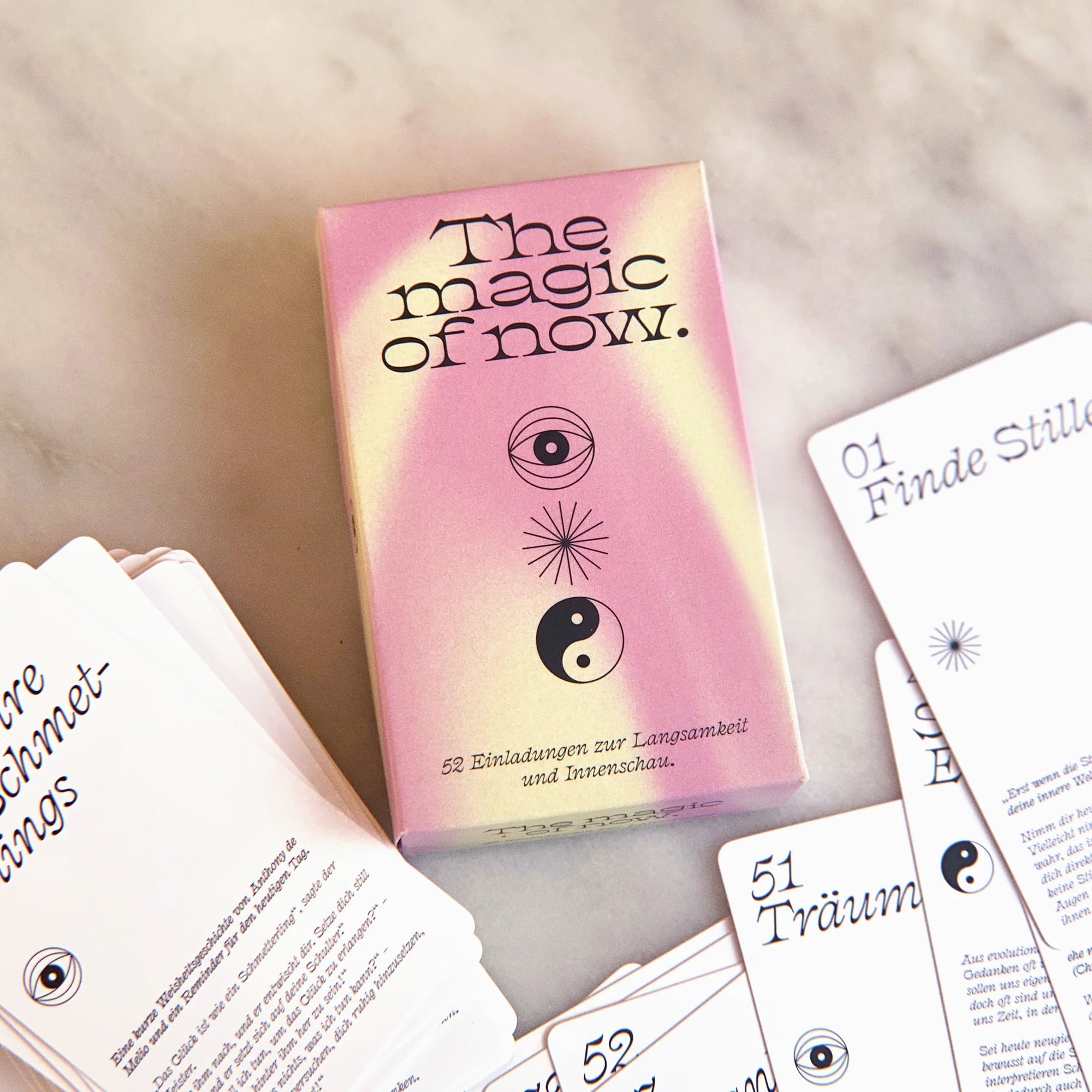 Eine pastellfarbene Kartenspielbox mit dem Titel The magic of now. mit einem Auge und einem Yin-Yang-Symbol, umgeben von mehreren teilweise sichtbaren Karten mit deutschem Text und Zahlen.