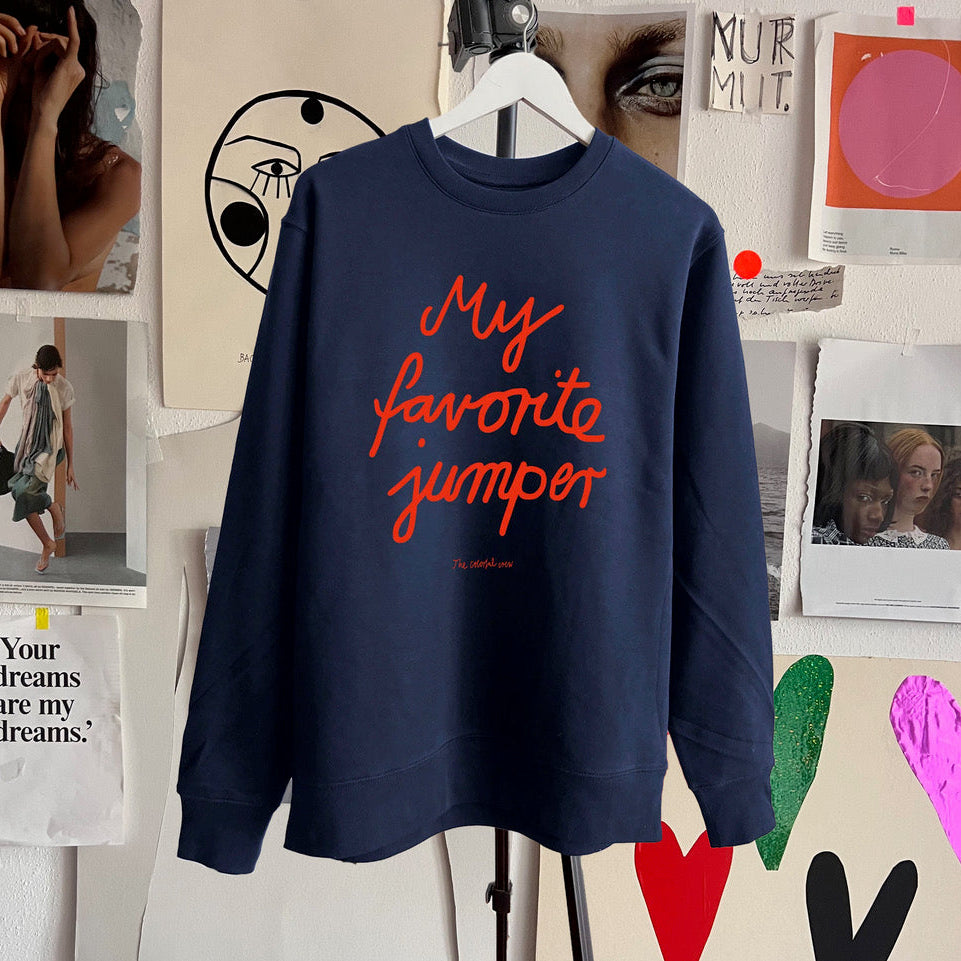 Ein marineblaues Sweatshirt mit dem Schriftzug "Mein Lieblingspullover" in fetter roter Schrift hängt an einem weißen Kleiderbügel. Der Hintergrund besteht aus einer vielseitigen Collage aus Kunstdrucken, Fotos und bunten ausgeschnittenen Herzen.