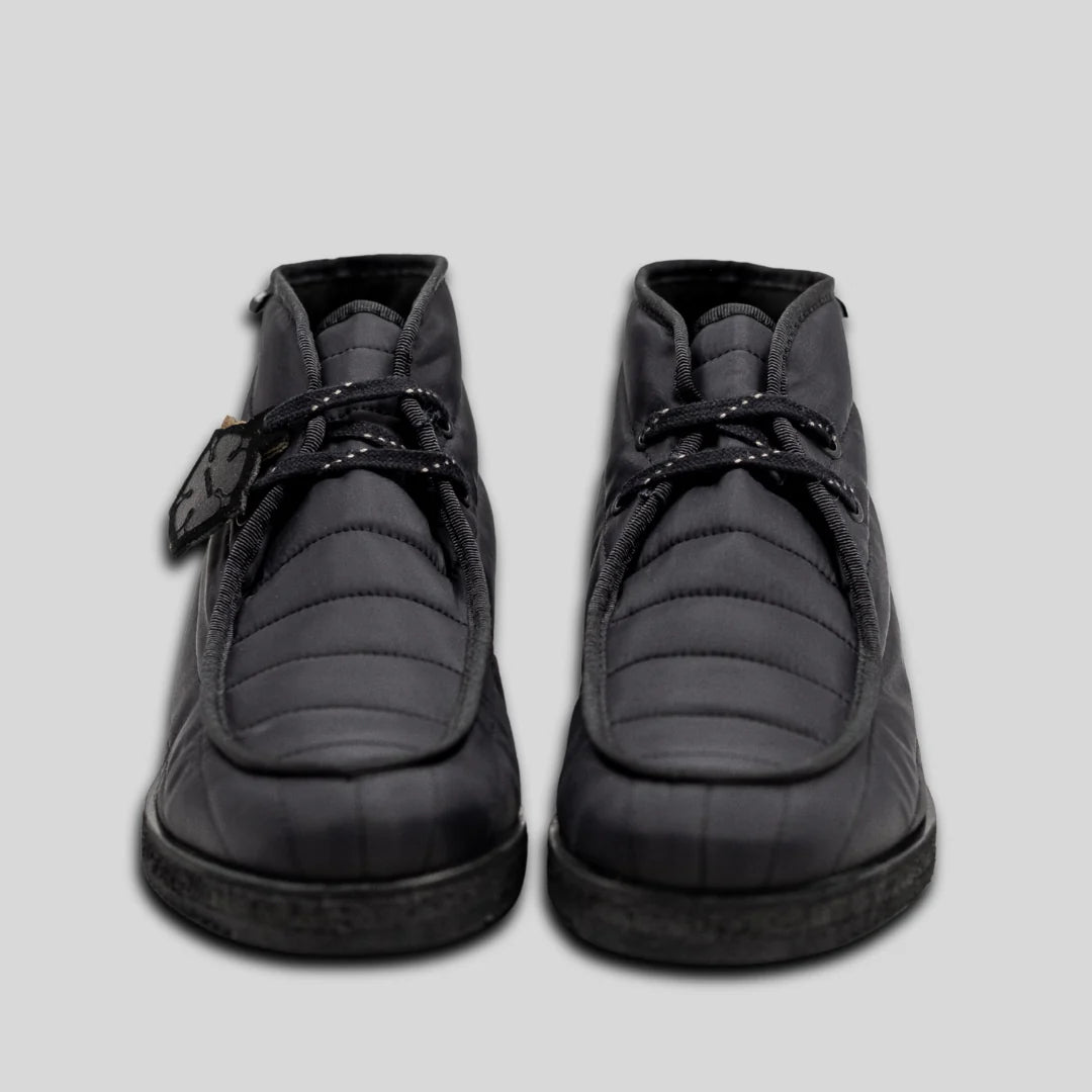 Ein Paar schwarze, gesteppte Schuhe mit schwarzen Schnürsenkeln, von vorne gesehen vor einem schlichten hellgrauen Hintergrund. An einem Schuh ist ein schwarzes Etikett am Schnürsenkel befestigt.