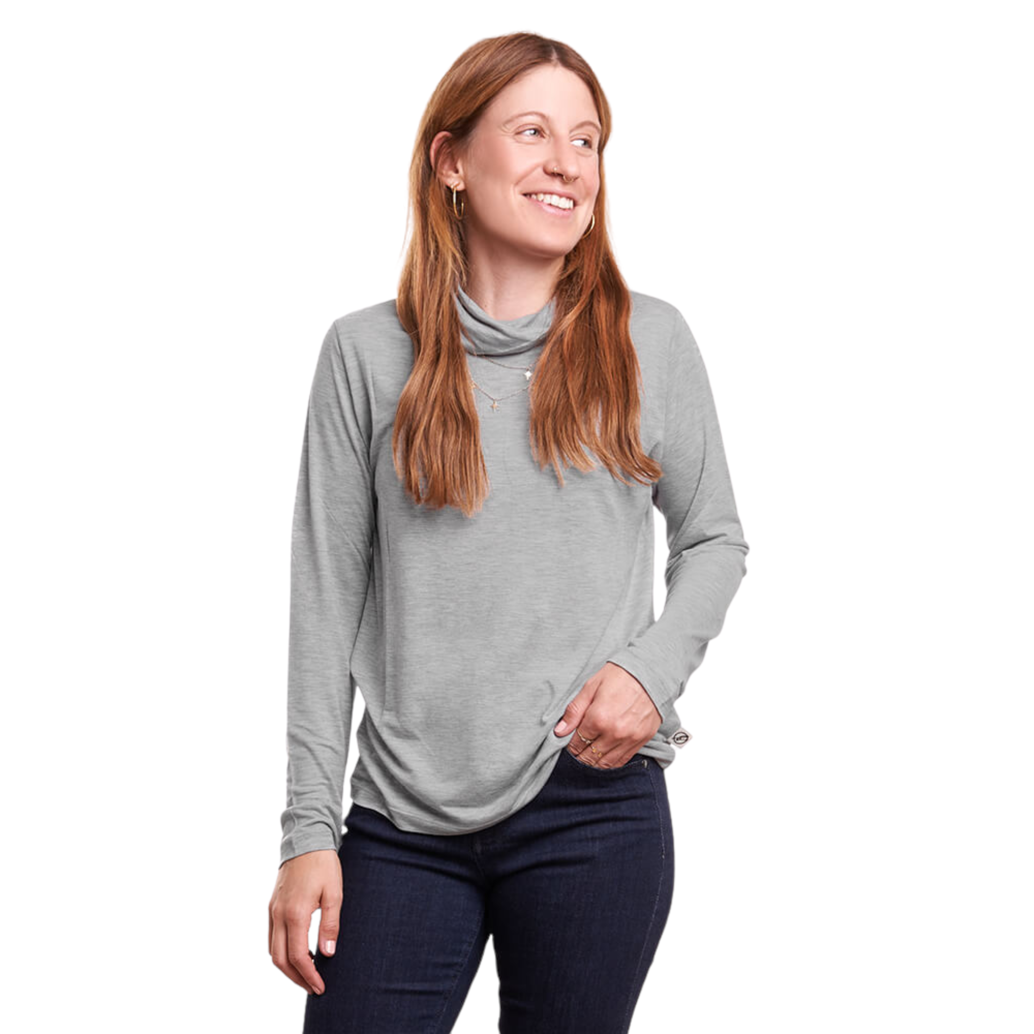 Frau in grauem Langarmshirt und dunklen Jeans lächelt mit der Hand in der Tasche auf weißem Hintergrund.