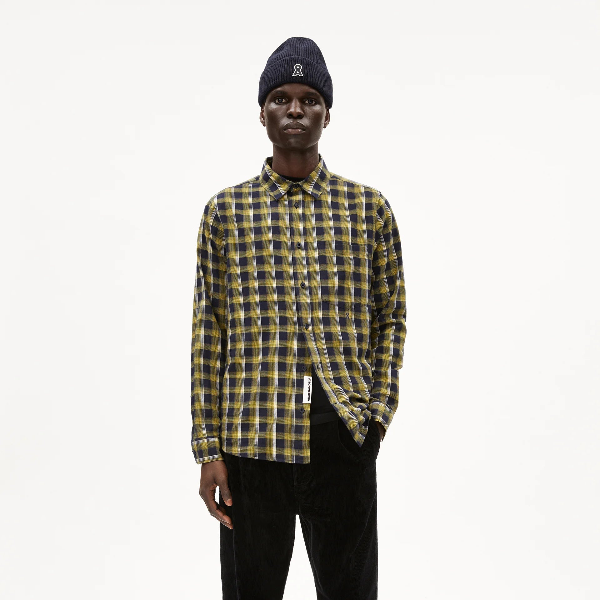 Eine Person, die ein marineblau-gelb kariertes Button-up-Hemd, eine schwarze Hose und eine schwarze Mütze trägt, steht vor einem schlichten weißen Hintergrund und blickt geradeaus, eine Hand in der Tasche.