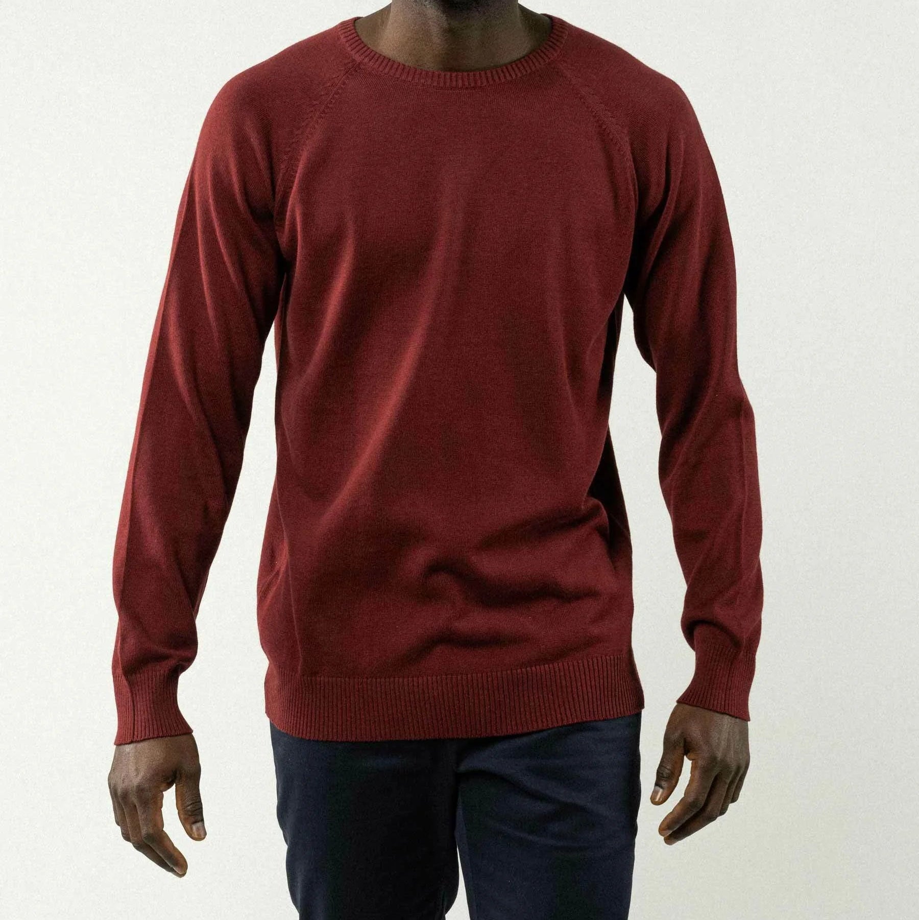 Ein Mann lächelt, während er vor einem schlichten hellen Hintergrund steht. Er trägt einen langärmeligen, dunkelroten Pullover und eine dunkle Hose. Seine Arme liegen entspannt an den Seiten.