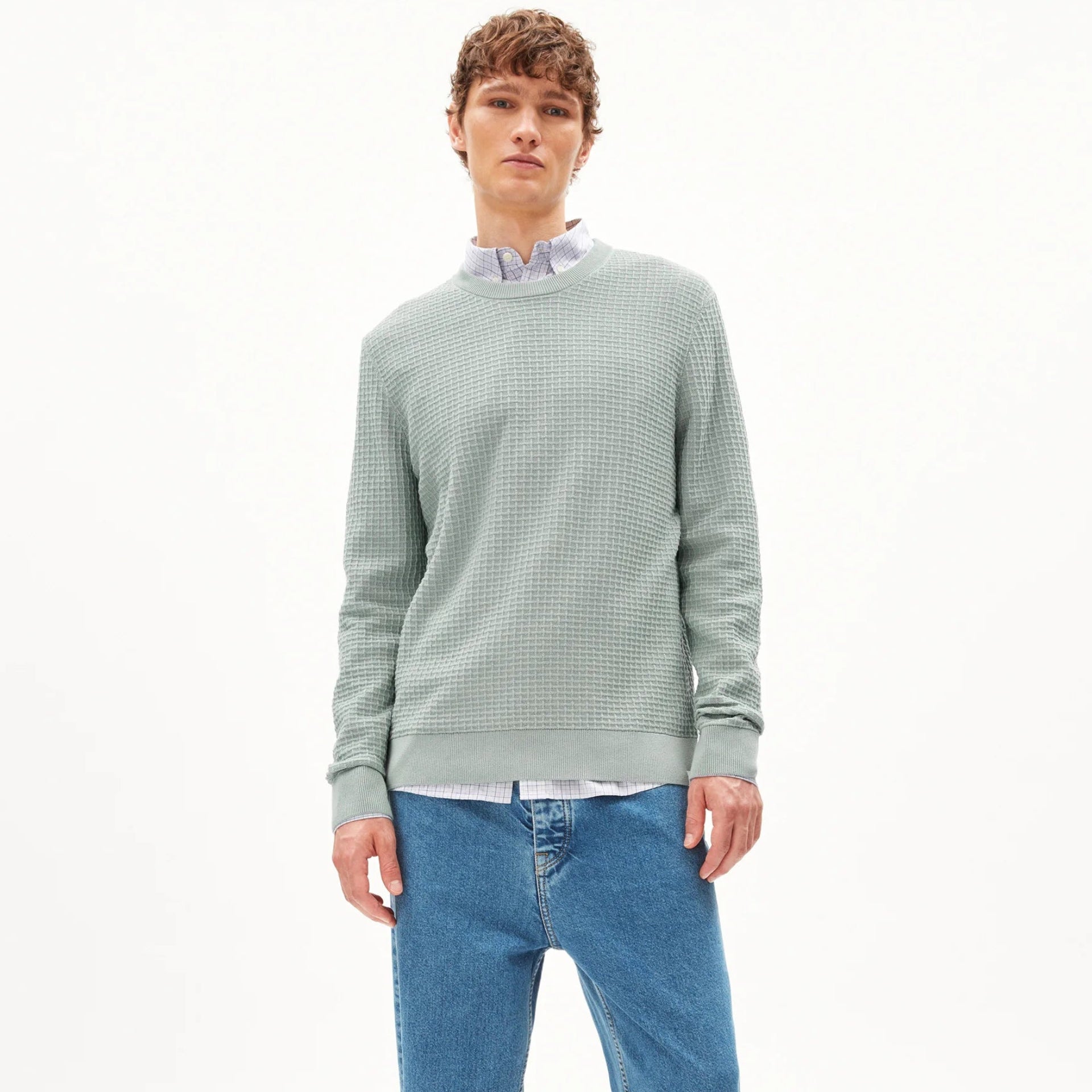 Ein junger Mann steht vor einem weißen Hintergrund. Er trägt einen hellgrünen strukturierten Pullover über einem weißen Hemd mit Kragen, weite blaue Jeans mit breiten Manschetten und schwarze Schuhe.