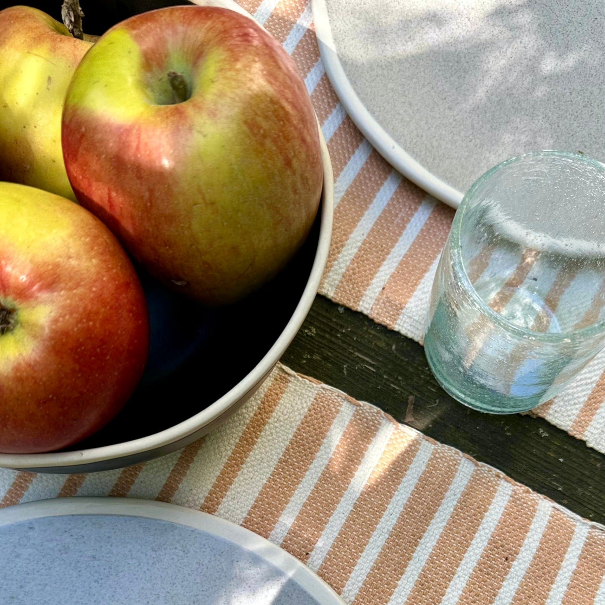 Schale mit Äpfeln neben Glas und Teller auf gestreifter Tischdecke im Sonnenlicht.