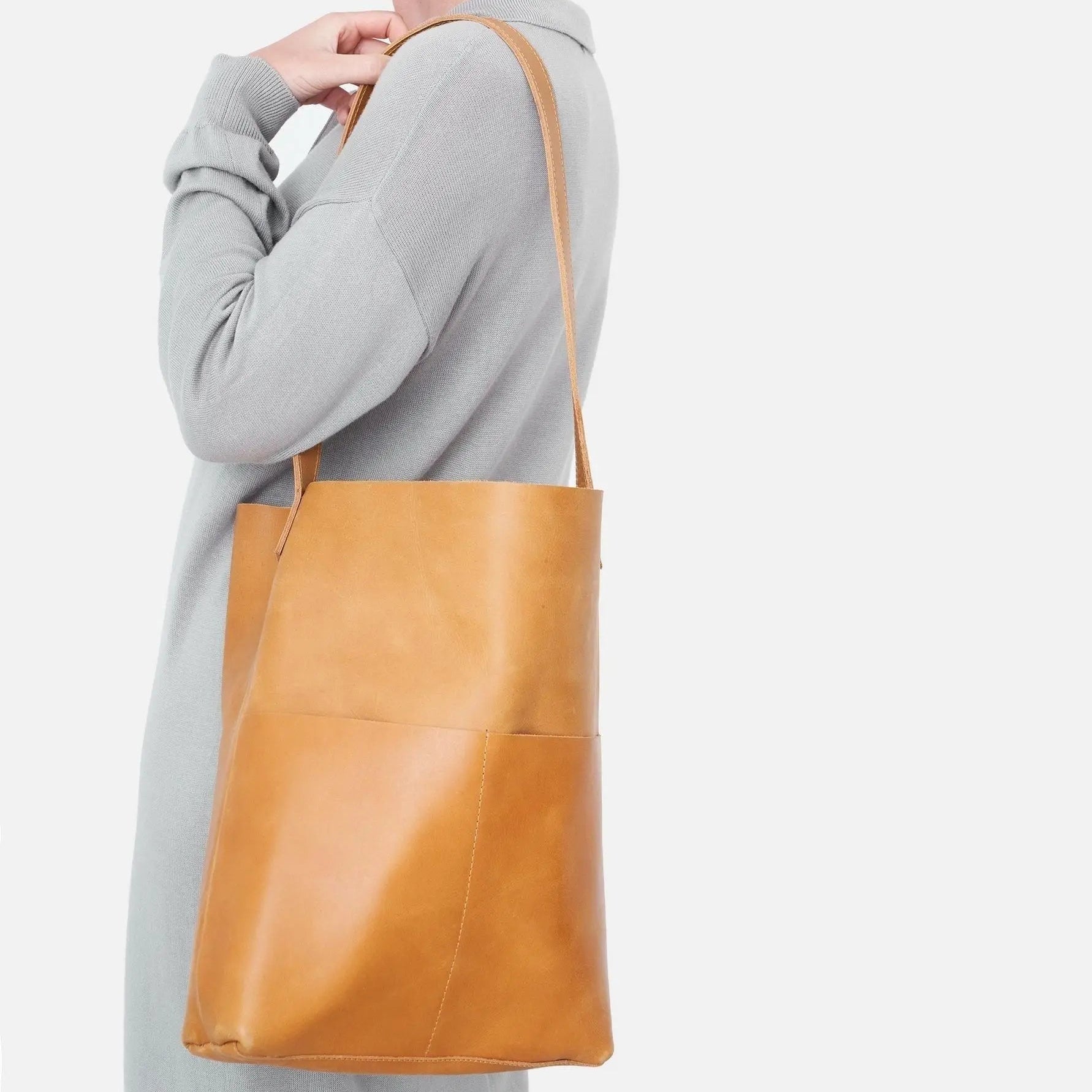 Eine Person, die einen hellgrauen Mantel trägt, hält eine hellbraune Lederumhängetasche mit dünnen Riemen. Die Tasche hängt an ihrer Seite vor einem schlichten weißen Hintergrund.