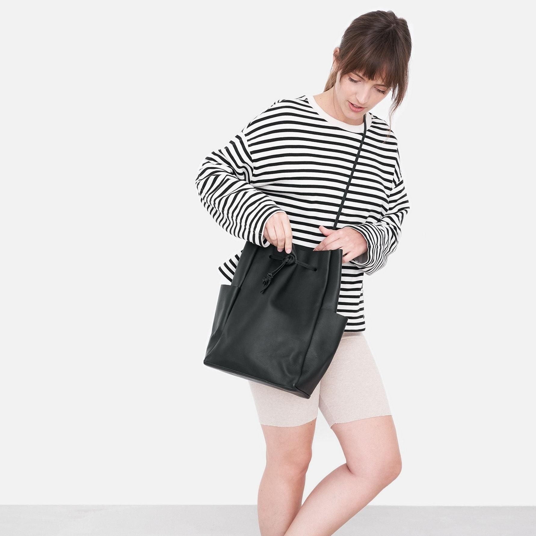 Eine Frau, die ein schwarz-weiß gestreiftes langärmeliges Hemd und beigefarbene Shorts trägt, schaut in eine große schwarze Umhängetasche vor einem schlichten weißen Hintergrund.