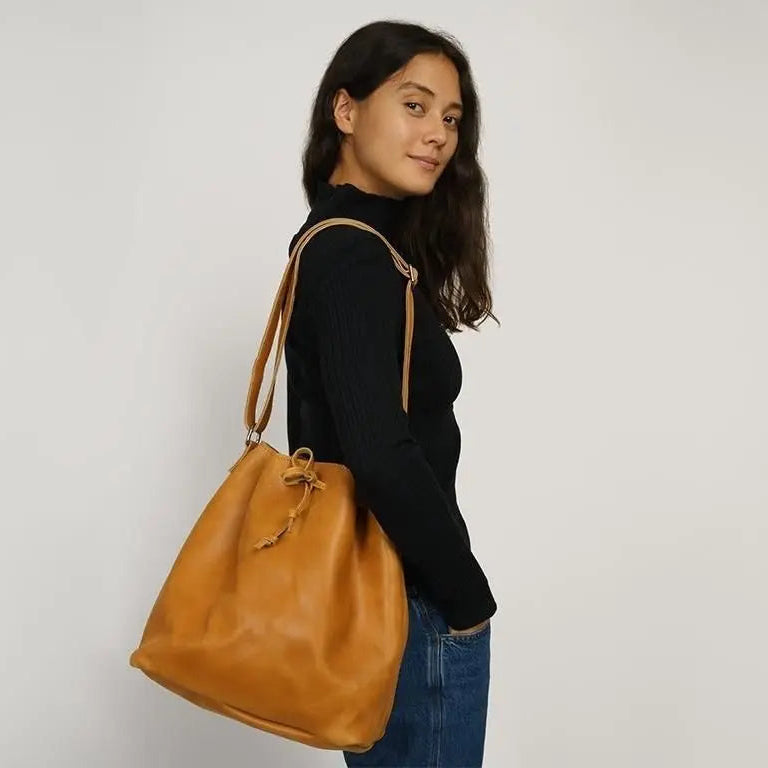 Eine Frau mit langen braunen Haaren, die einen schwarzen Rollkragenpullover und blaue Jeans trägt, steht seitlich und blickt in die Kamera. Sie trägt eine große hellbraune Lederumhängetasche auf ihrem Arm vor einem schlichten hellen Hintergrund.