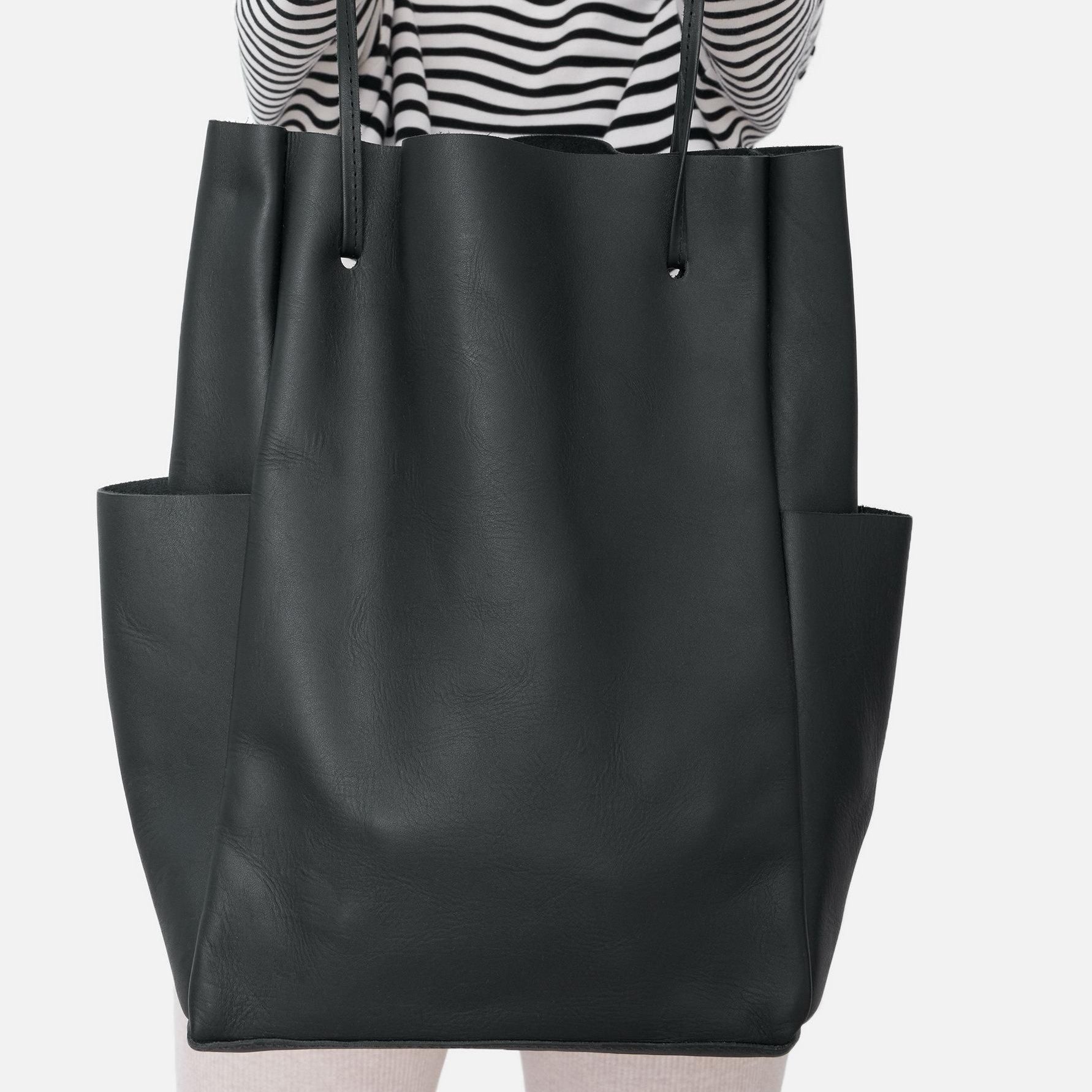Eine Person, die ein schwarz-weiß gestreiftes Hemd trägt, hält eine große schwarze Ledertasche mit zwei Seitentaschen, die vor einem schlichten weißen Hintergrund gezeigt wird.