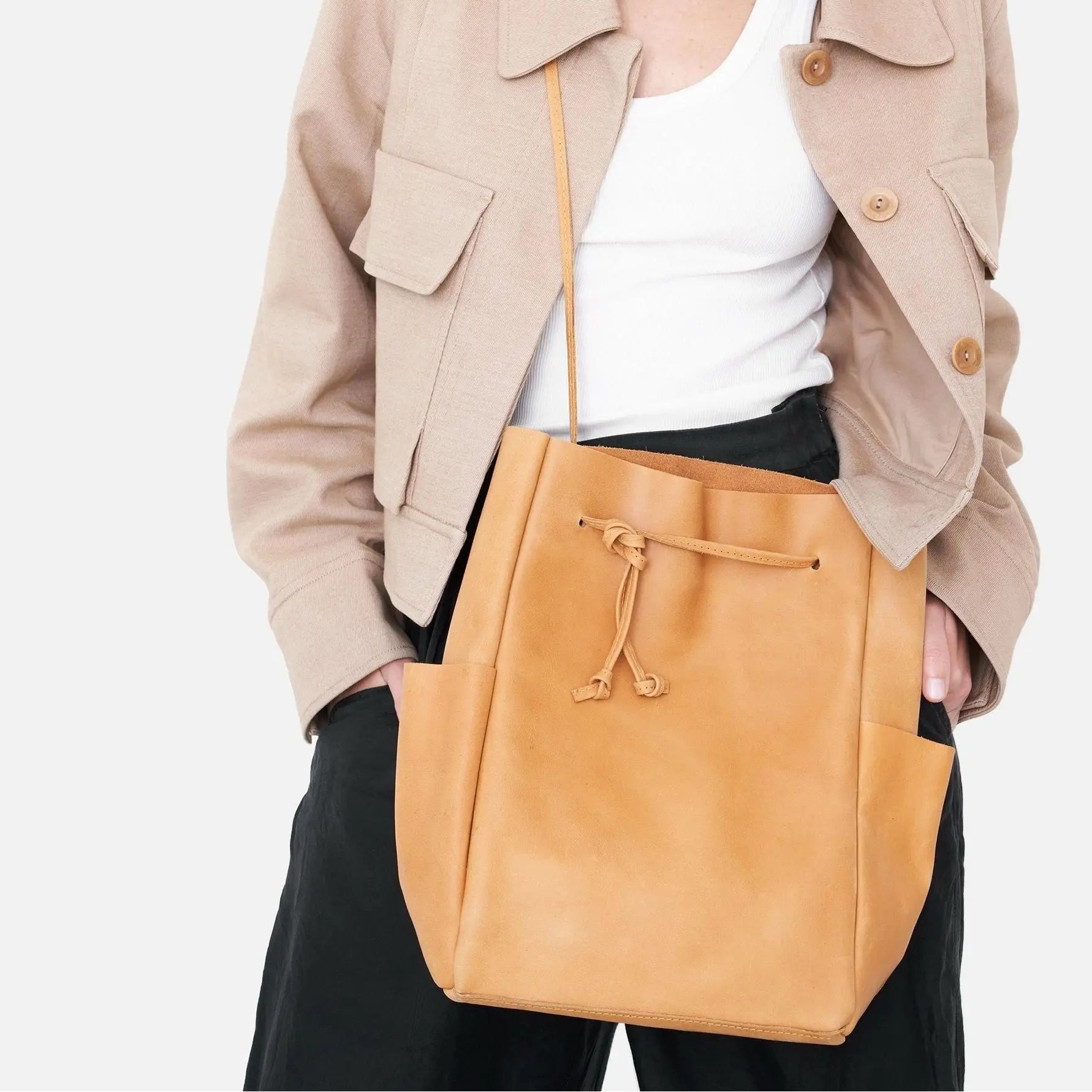 Eine Person, die eine beige Jacke, ein weißes Oberteil und eine schwarze Hose trägt, hält eine hellbraune Lederumhängetasche mit Vordertaschen und einem Kordelzugverschluss. Der Hintergrund ist einfarbig und hell.