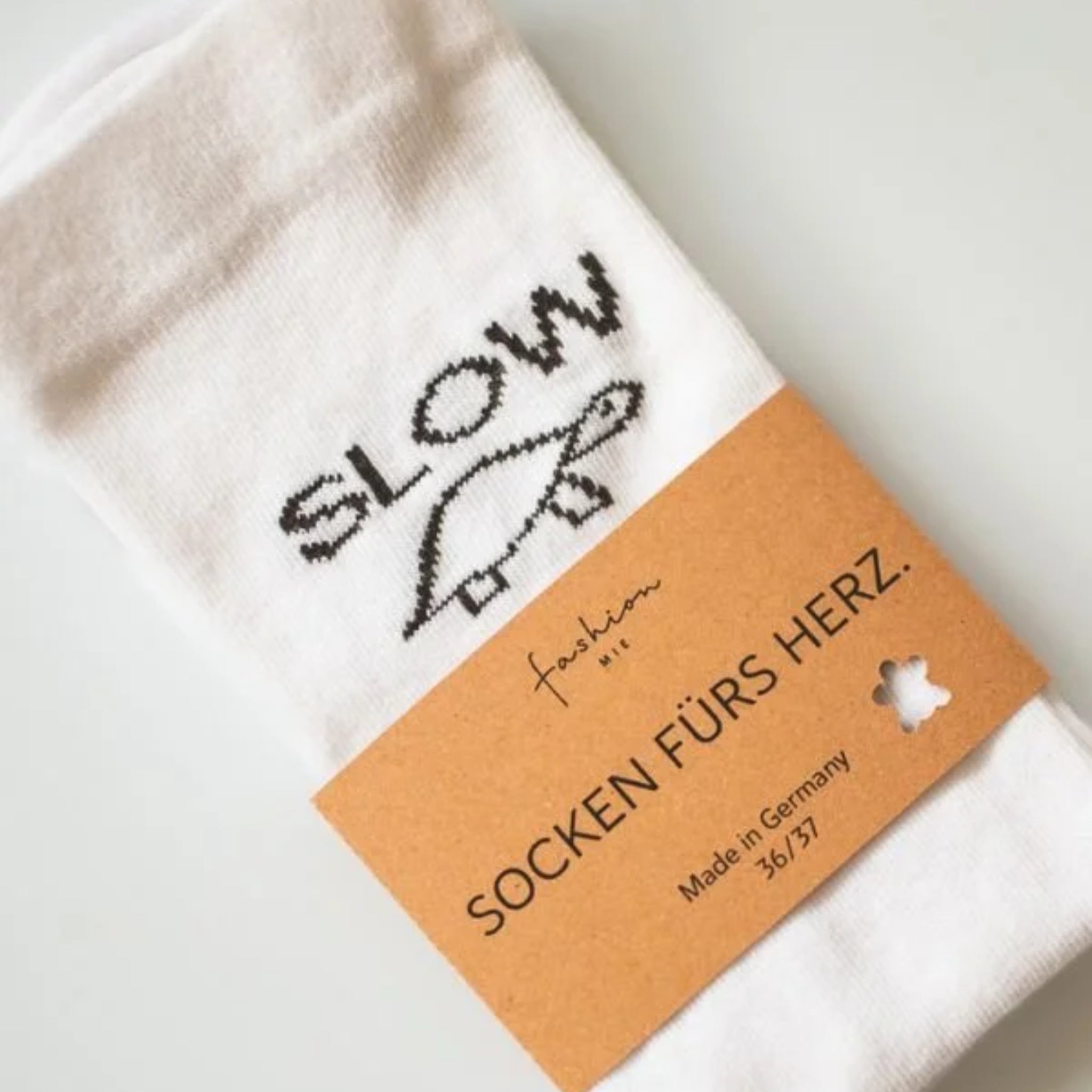 Ein Paar weiße Socken mit aufgestickter Schildkröte und dem Wort SLOW, umhüllt von einem braunen Etikett mit der Aufschrift SOCKEN FÜRS HERZ. Hergestellt in Deutschland 36/37.