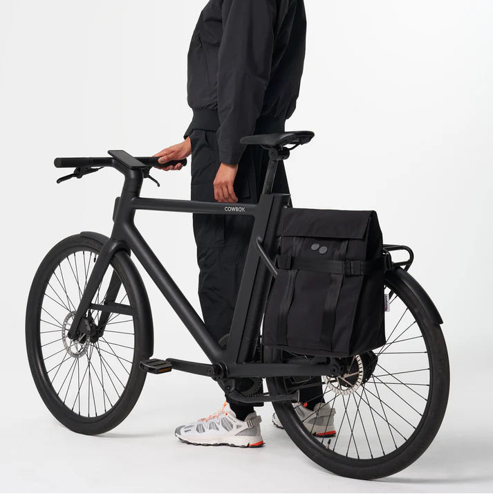 Eine schwarz gekleidete Person steht neben einem mattschwarzen Fahrrad mit einer großen schwarzen Gepäckträgertasche auf dem Gepäckträger. Der Hintergrund ist einfarbig weiß.