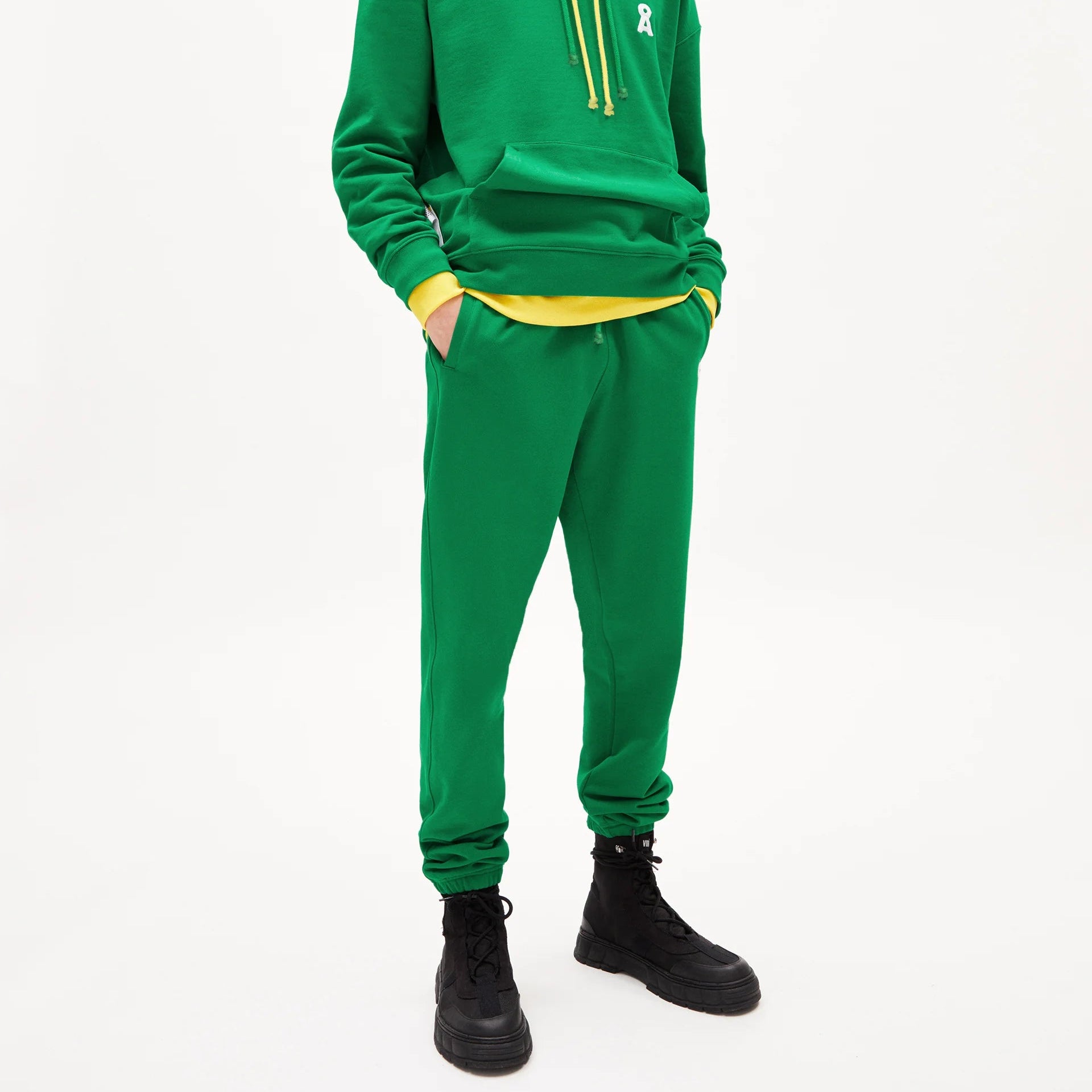 Eine Person mit langen blonden Haaren trägt einen grünen Kapuzenpulli mit gelben Kordelzügen über einem gelben Kapuzenpulli, eine passende grüne Jogginghose und schwarze Stiefel und steht vor einem schlichten weißen Hintergrund.