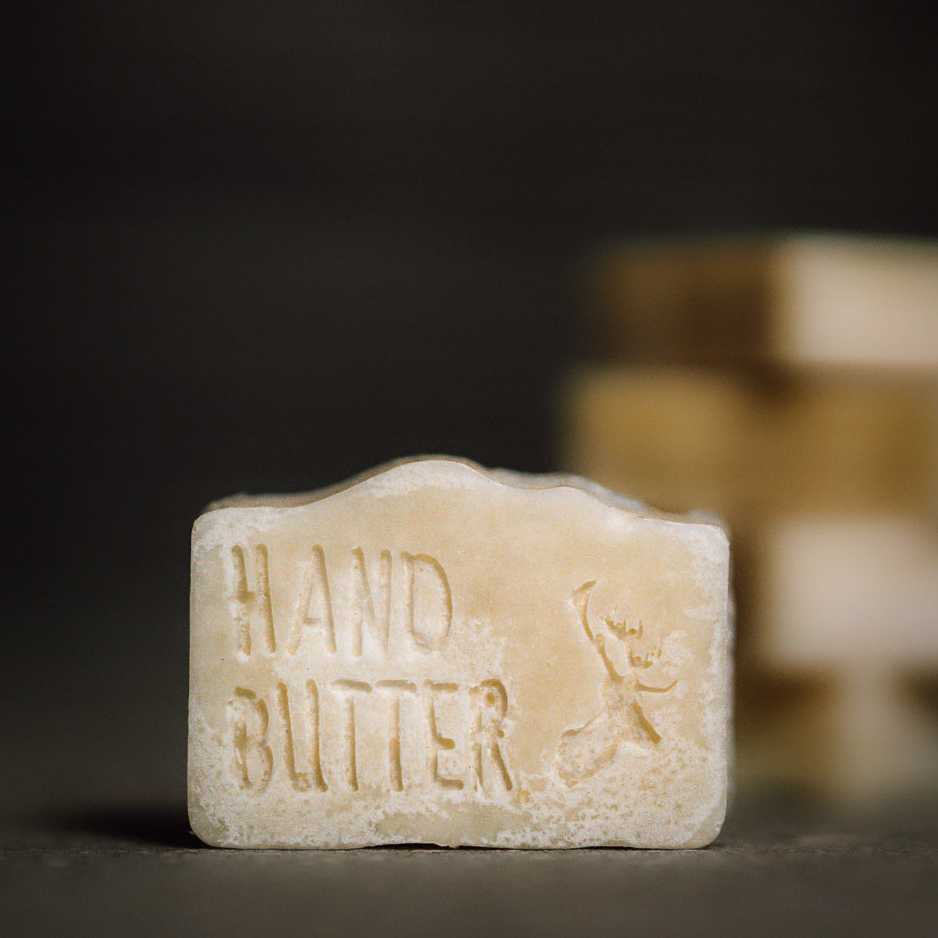 Ein rechteckiges Seifenstück mit der Aufschrift "HAND BUTTER" und einer kleinen Figur mit ausgestreckten Armen, die eingraviert ist. Der Hintergrund ist unscharf mit gestapelten Seifenstücken.