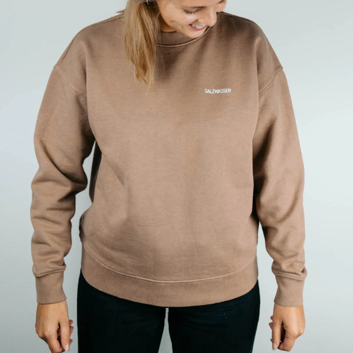Eine Person, die ein lockeres, braunes Sweatshirt trägt, auf dessen Brust in weißer Farbe das Wort SALZWASSER" aufgestickt ist, steht vor einem schlichten, hellen Hintergrund und schaut mit einem leichten Lächeln nach unten.