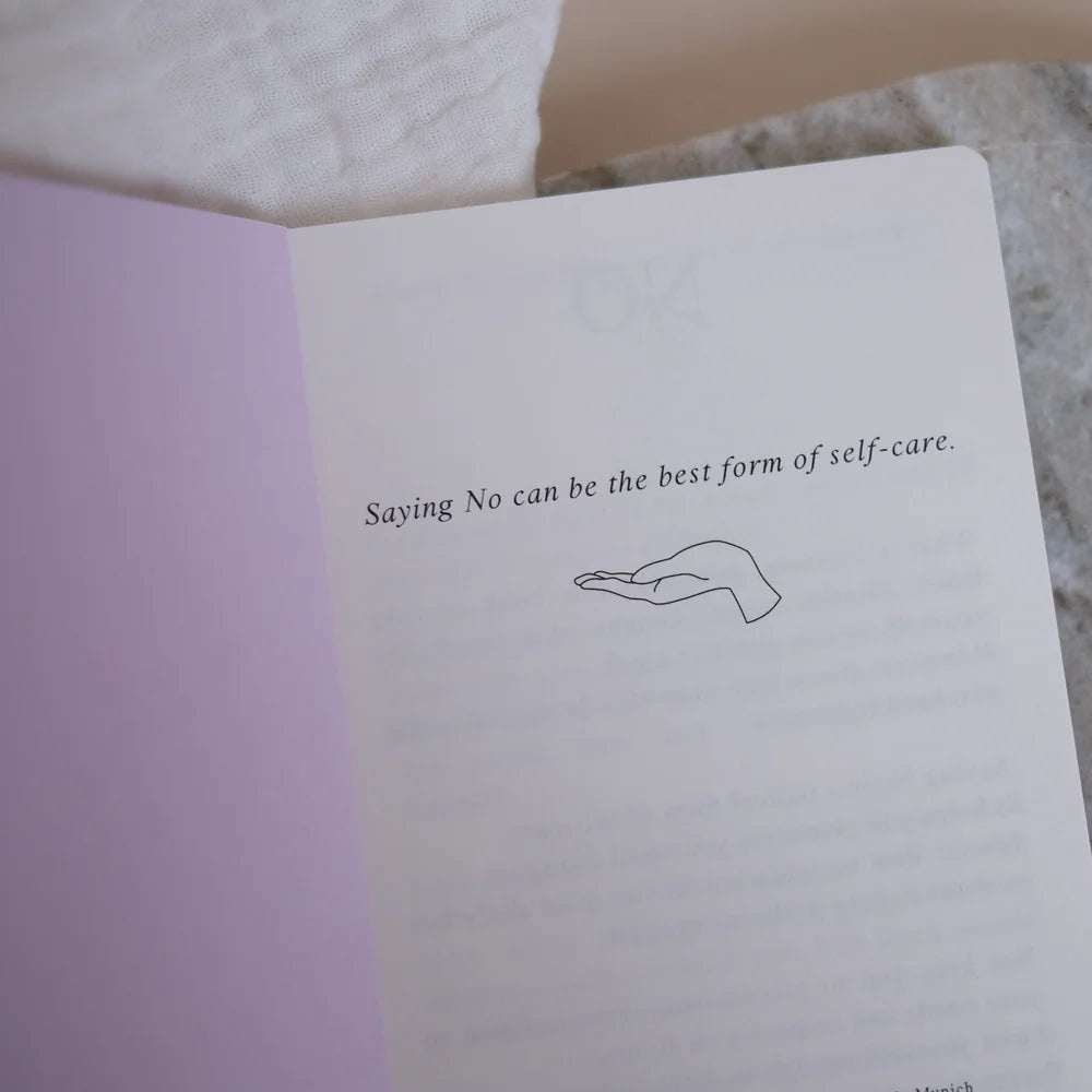 Ein aufgeschlagenes Buch mit dem Text Saying No can be the best form of self-care" (Nein sagen kann die beste Form der Selbstfürsorge sein) liegt über einer einfachen Strichzeichnung einer offenen Hand; daneben ruht ein Paar goldener Ohrringe auf einer Marmorfläche.