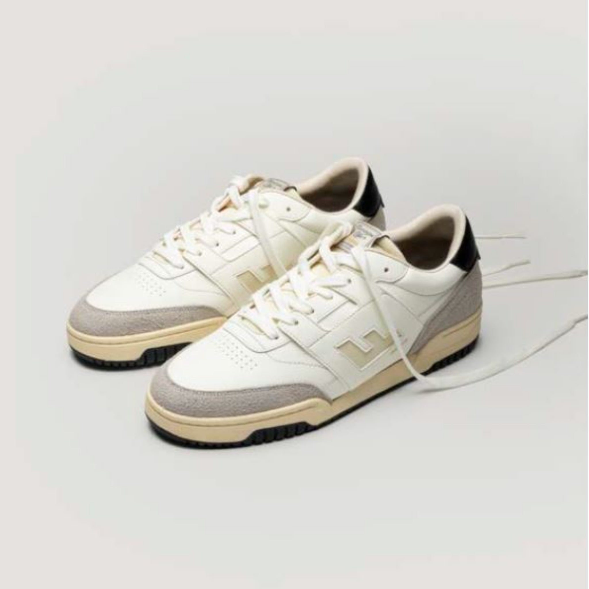 Ein Paar weiße Turnschuhe mit beigen und hellgrauen Akzenten, cremefarbenen Sohlen und schwarzen Details an den Absätzen. Die Schuhe stehen nebeneinander auf einem schlichten hellen Hintergrund, die Schnürsenkel sind geöffnet.