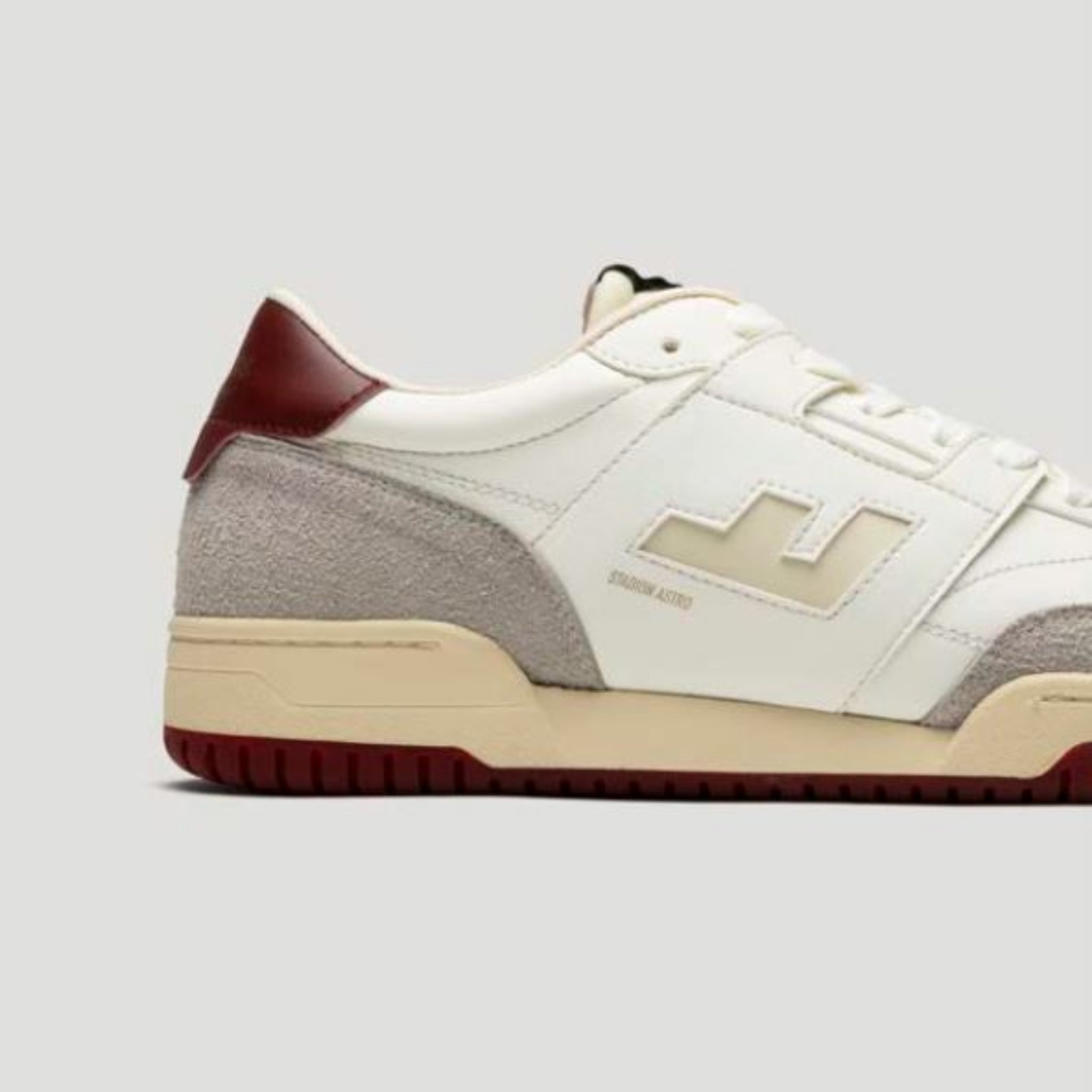 Ein weißer Sneaker mit beigen und grauen Akzenten, einer roten Fersenlasche und einer strukturierten Sohle. Der Schuh hat ein auffälliges geometrisches Muster an der Seite und ist vor einem schlichten grauen Hintergrund fotografiert.