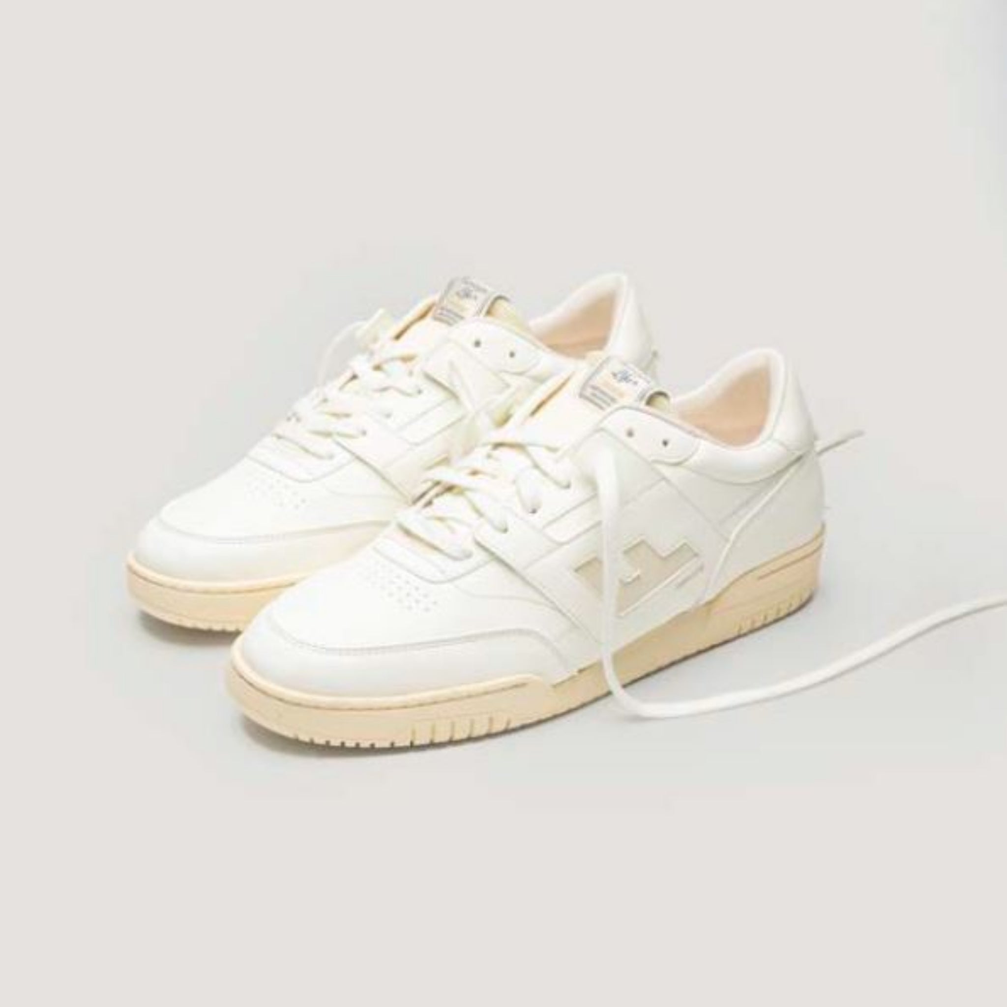 Ein Paar cremeweiße Turnschuhe mit cremefarbener Sohle und Schnürsenkeln, mit seitlichem Logo und minimalistischem Design auf hellgrauem Hintergrund.