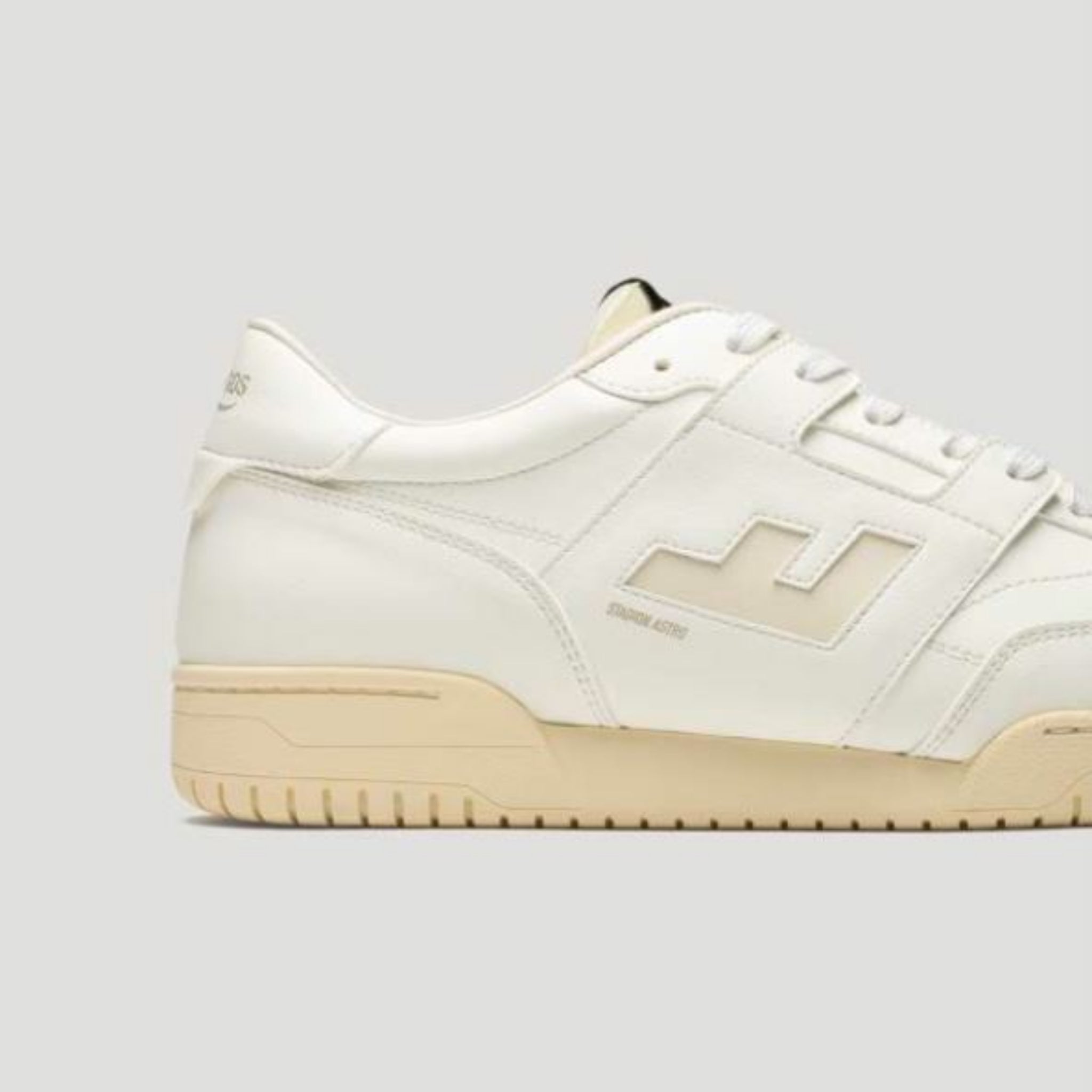 Ein cremefarbener Sneaker mit beiger Sohle, einem dezenten geometrischen Logo an der Seite und weißen Schnürsenkeln, die vor einem schlichten hellgrauen Hintergrund stehen.