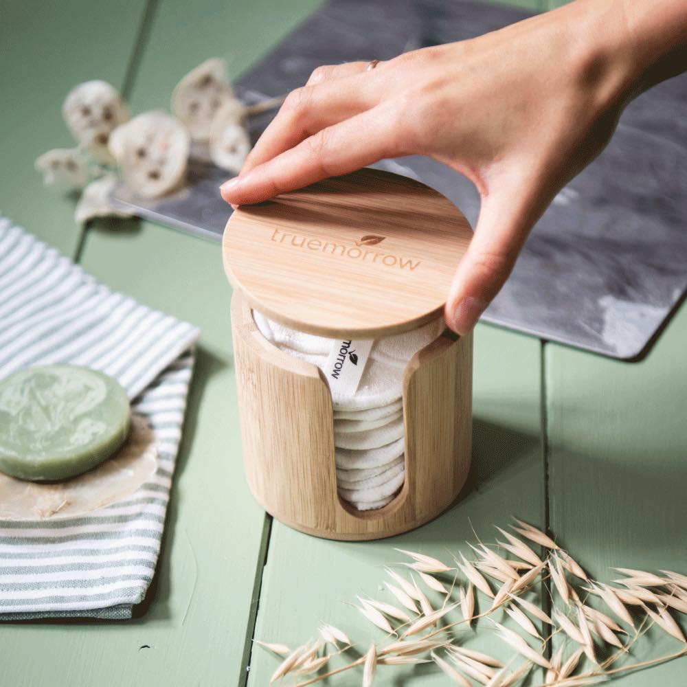 Eine Hand öffnet einen Bambusbehälter mit runden Baumwollpads auf einem grünen Holztisch.