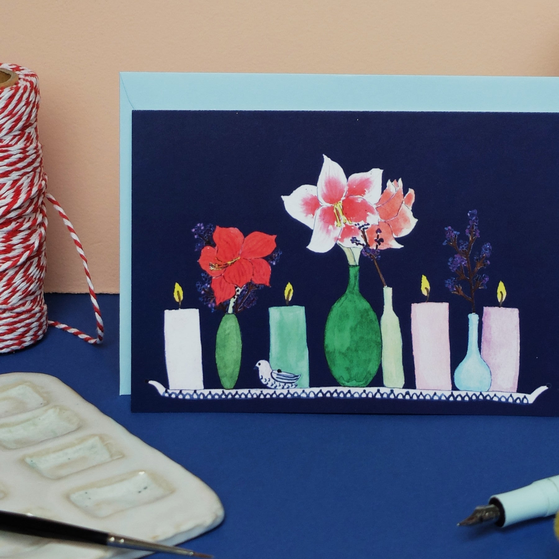 Eine Grußkarte mit Abbildungen von Vasen mit roten Blumen und brennenden Kerzen, die auf einem Tablett stehen. Rote und weiße Schnüre, eine Aquarellpalette und ein Füllfederhalter liegen in der Nähe auf einer blauen Fläche.