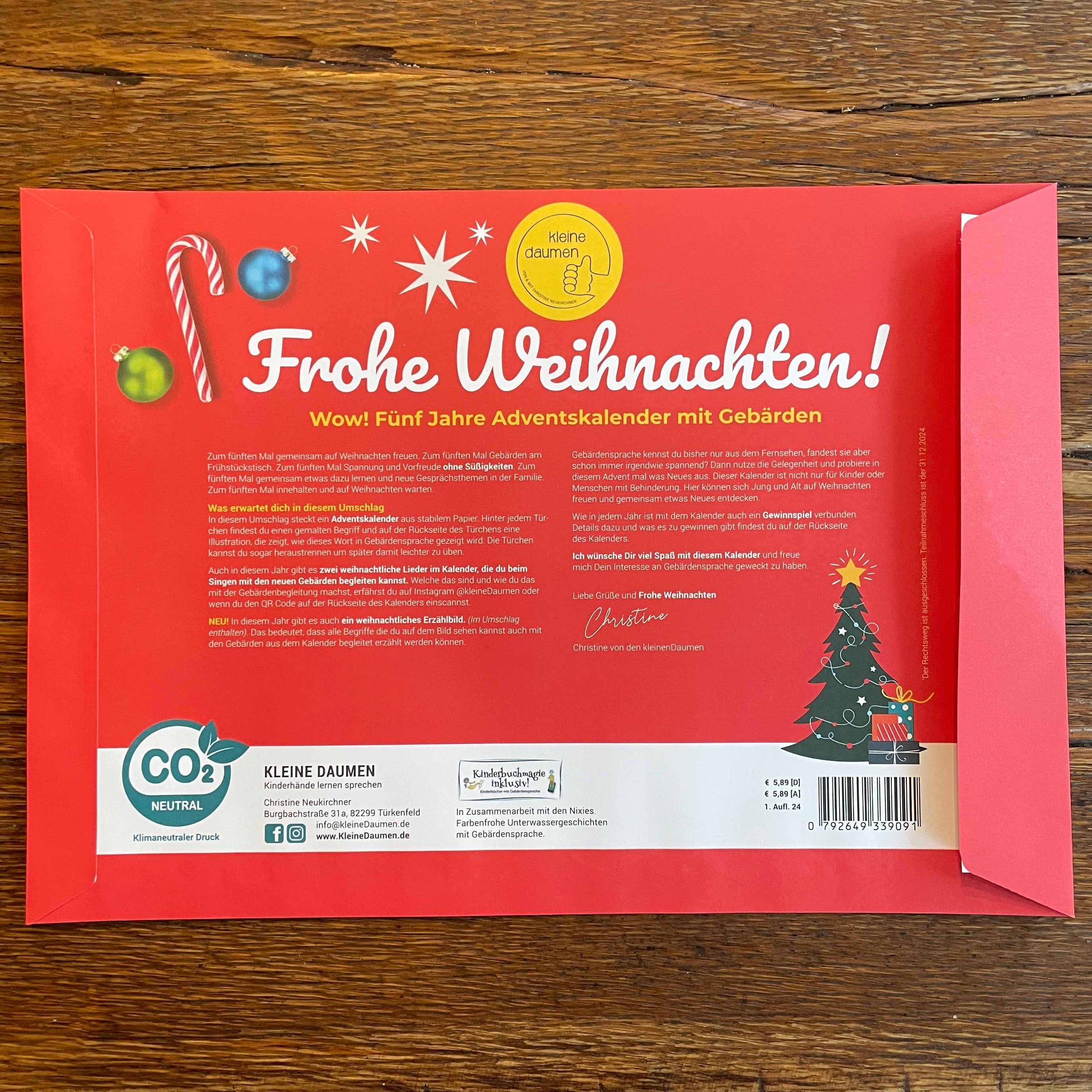 Roter Umschlag mit dem deutschen Text Frohe Weihnachten! und festlichen Grafiken wie einer Zuckerstange, einem grünen Ornament und einem Weihnachtsbaum. Es gibt Logos, einen Strichcode und weiße und gelbe Schrift über einen Adventskalender.