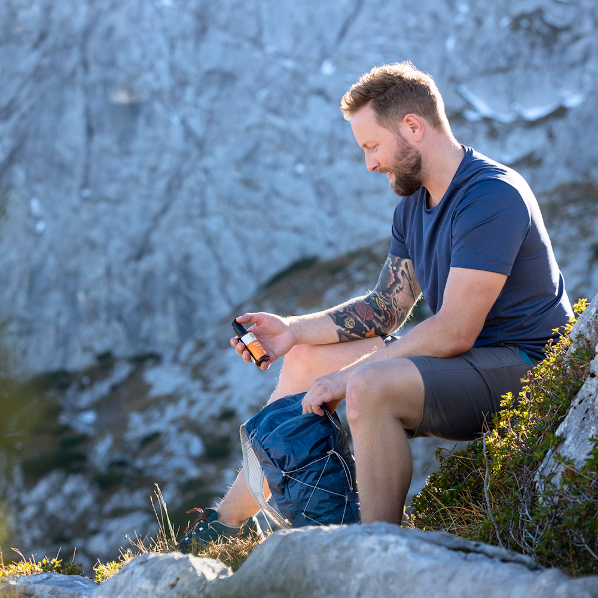 Ein Mann mit Bart und Tätowierungen sitzt im Freien auf einem Felsvorsprung, hält eine bernsteinfarbene Flasche in der einen Hand und einen blauen Rucksack auf dem Schoß, während im Hintergrund eine Berglandschaft zu sehen ist.