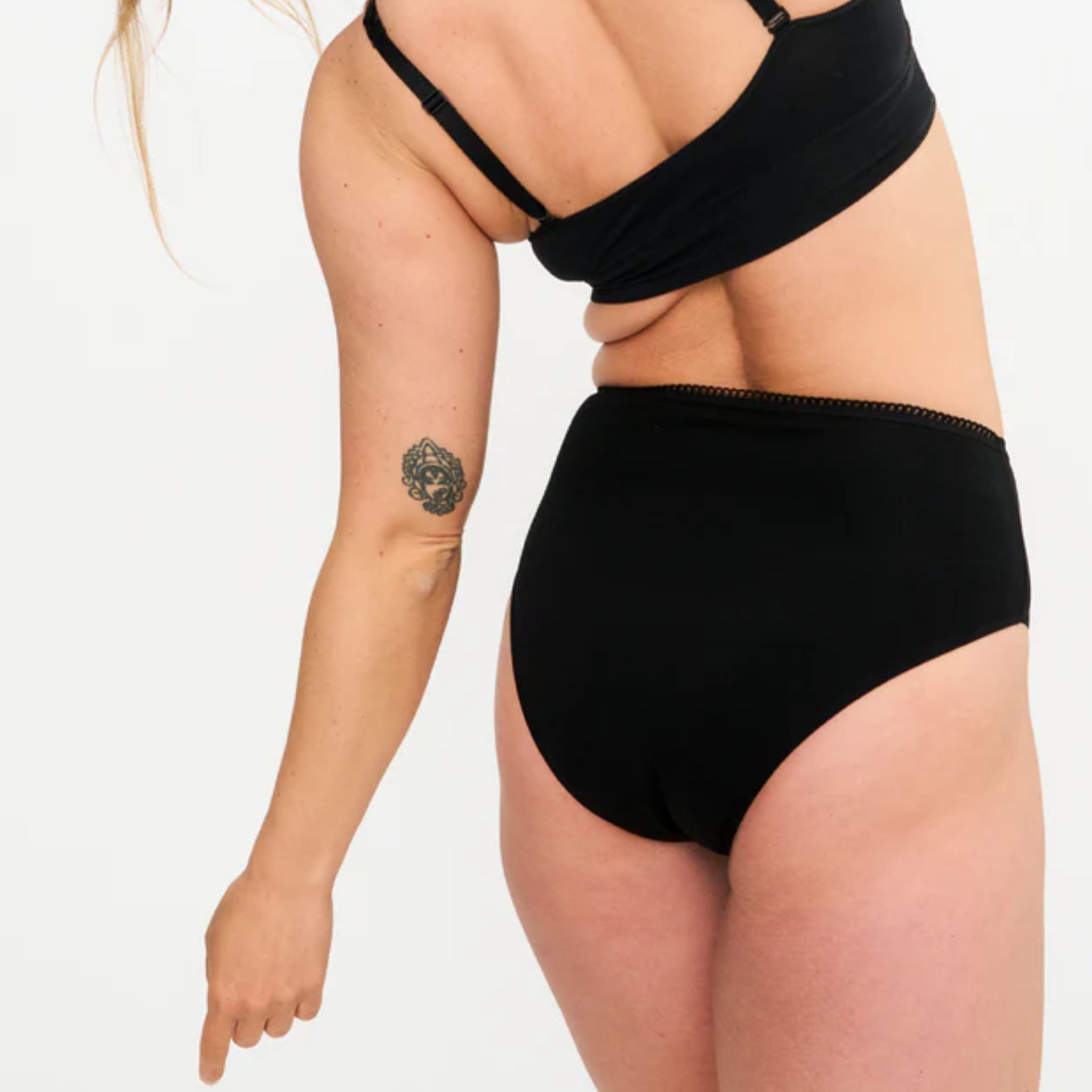 Eine Person, die einen schwarzen BH und schwarze Unterwäsche mit hoher Taille trägt, steht vor einem einfarbigen Hintergrund, wobei ihr Oberkörper und ihr Arm sichtbar sind. Die Person hat eine Tätowierung auf dem Oberarm und deutet mit der linken Hand nach unten.