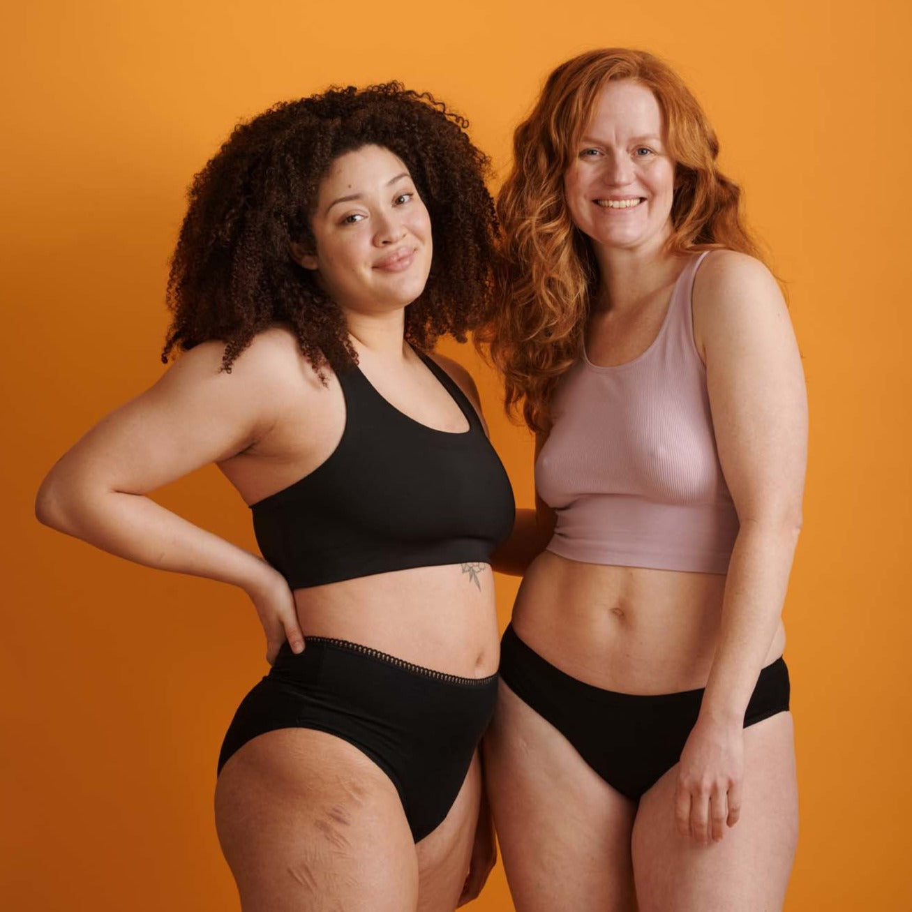 Zwei Frauen in Unterwäsche stehen vor einem orangefarbenen Hintergrund und lächeln in die Kamera.