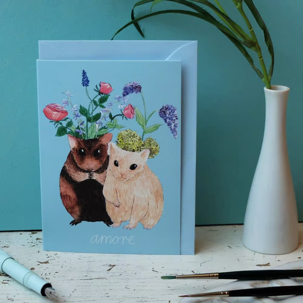 Eine Grußkarte mit zwei illustrierten Hamstern, aus deren Köpfen Blumen und Grünzeug wachsen, vor einem hellblauen Hintergrund. Darunter steht das Wort amore geschrieben. In der Nähe befinden sich eine Vase und Malutensilien.
