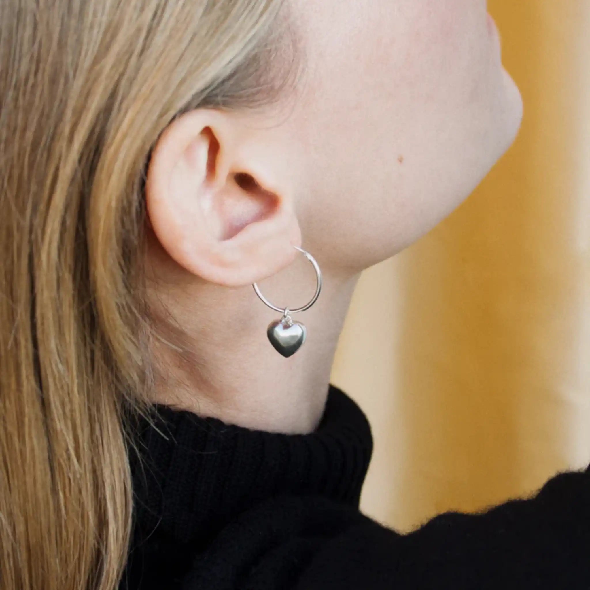 Nahaufnahme einer Person mit blondem Haar, die einen silbernen Ohrring mit einem herzförmigen Anhänger trägt, bekleidet mit einem schwarzen Rollkragenpullover, die leicht von der Kamera wegschaut.