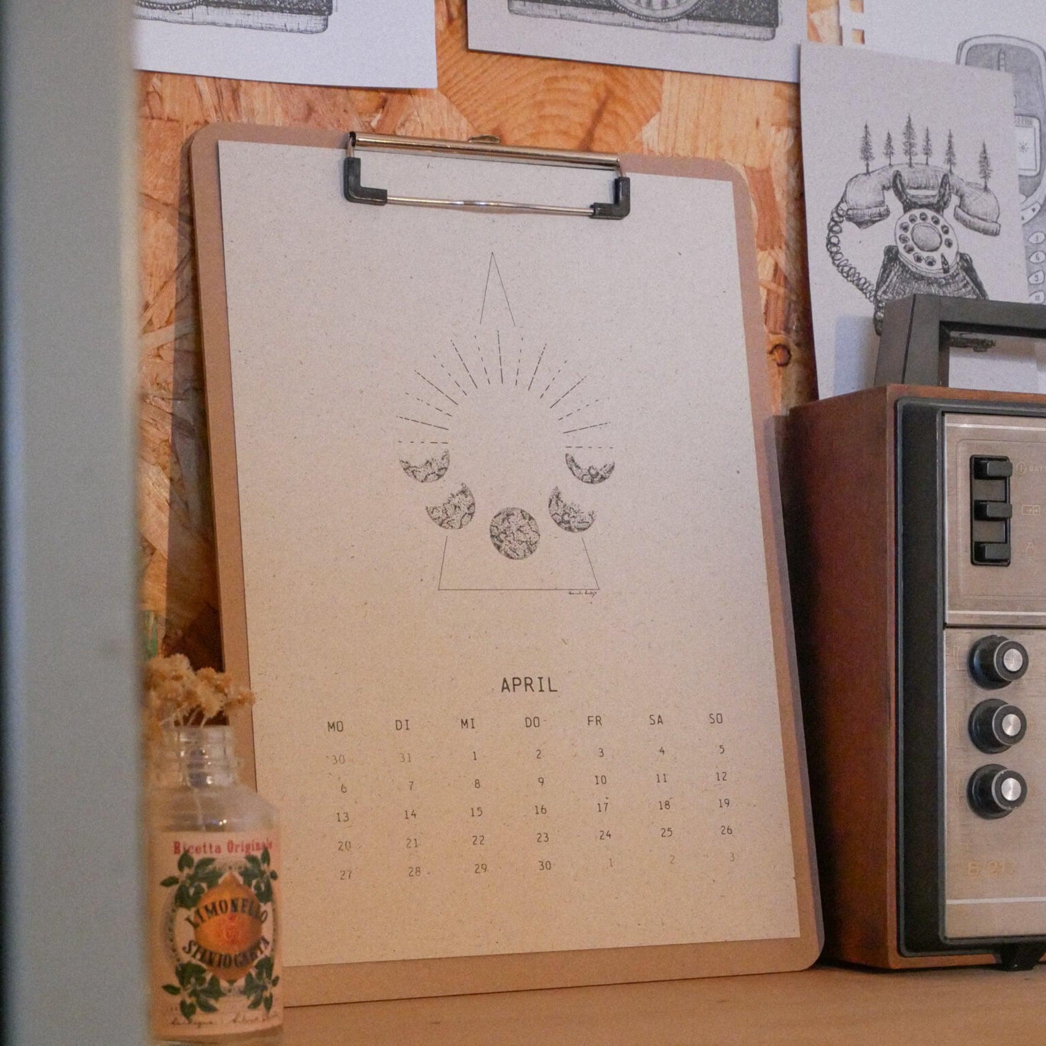Auf einem Klemmbrett liegt ein April-Kalenderblatt mit Mondphasen und einem geometrischen Muster. In der Nähe stehen ein altes Radio, eine kleine Glasflasche und Schwarz-Weiß-Zeichnungen, die an eine Holzwand gepinnt sind.