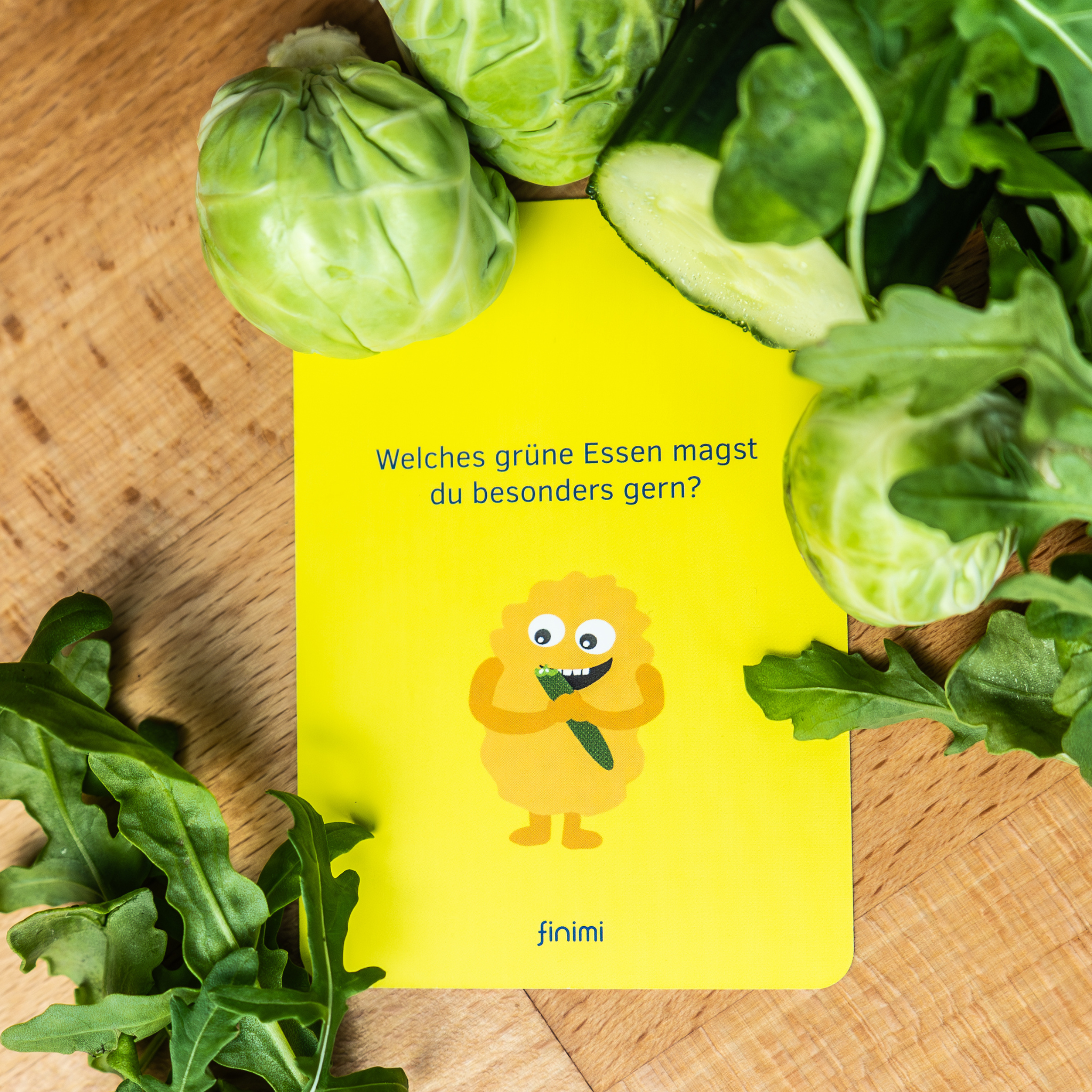 Eine gelbe Karte mit einer Zeichentrickfigur, die eine Gurke hält, umgeben von grünem Gemüse wie Rucola, Rosenkohl und Gurkenscheiben. Die Karte fragt auf Deutsch: Welches grüne Essen magst du besonders gerne?.