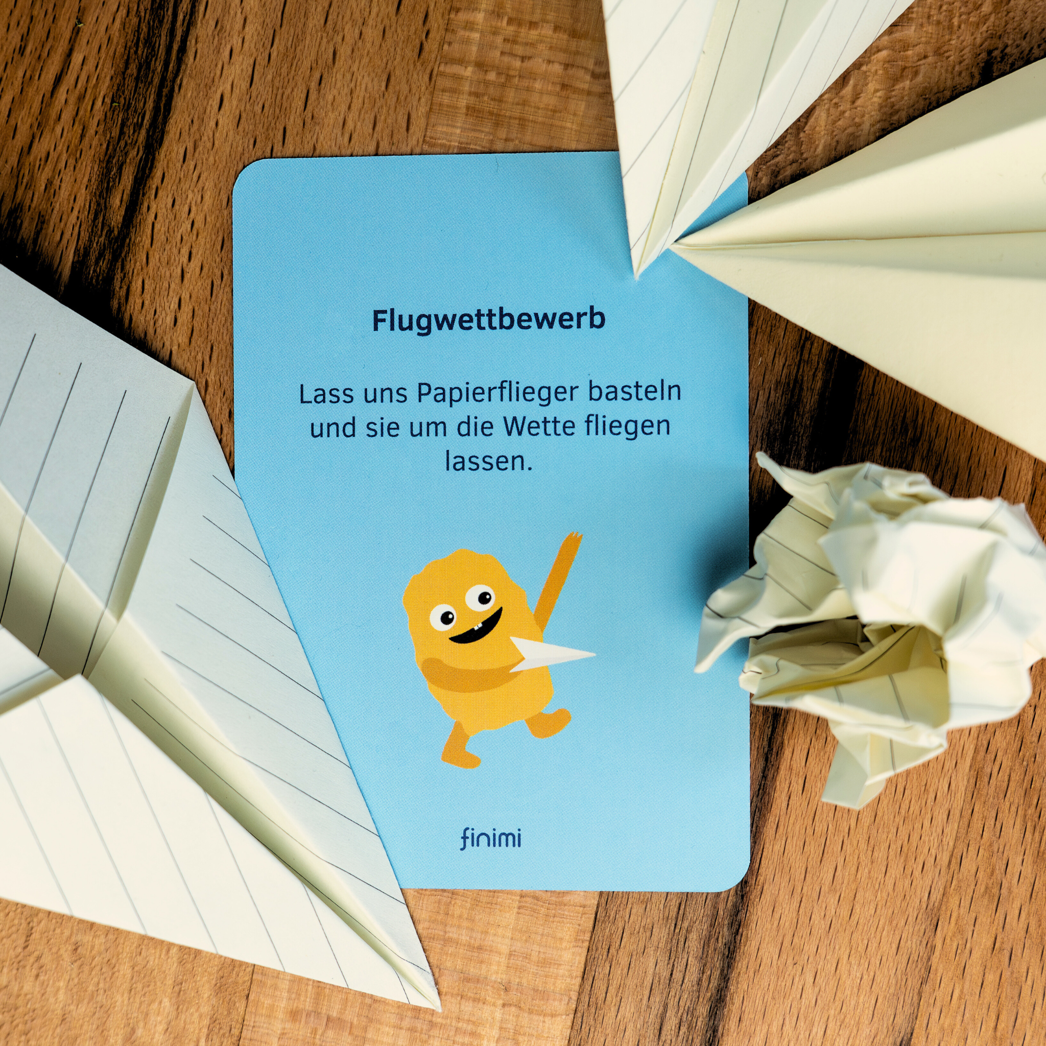 Eine blaue Karte mit deutschem Text und einer lächelnden Zeichentrickfigur, die ein Papierflugzeug hält, liegt auf einer hölzernen Fläche, umgeben von Papierflugzeugen und einem zerknüllten Papierball.