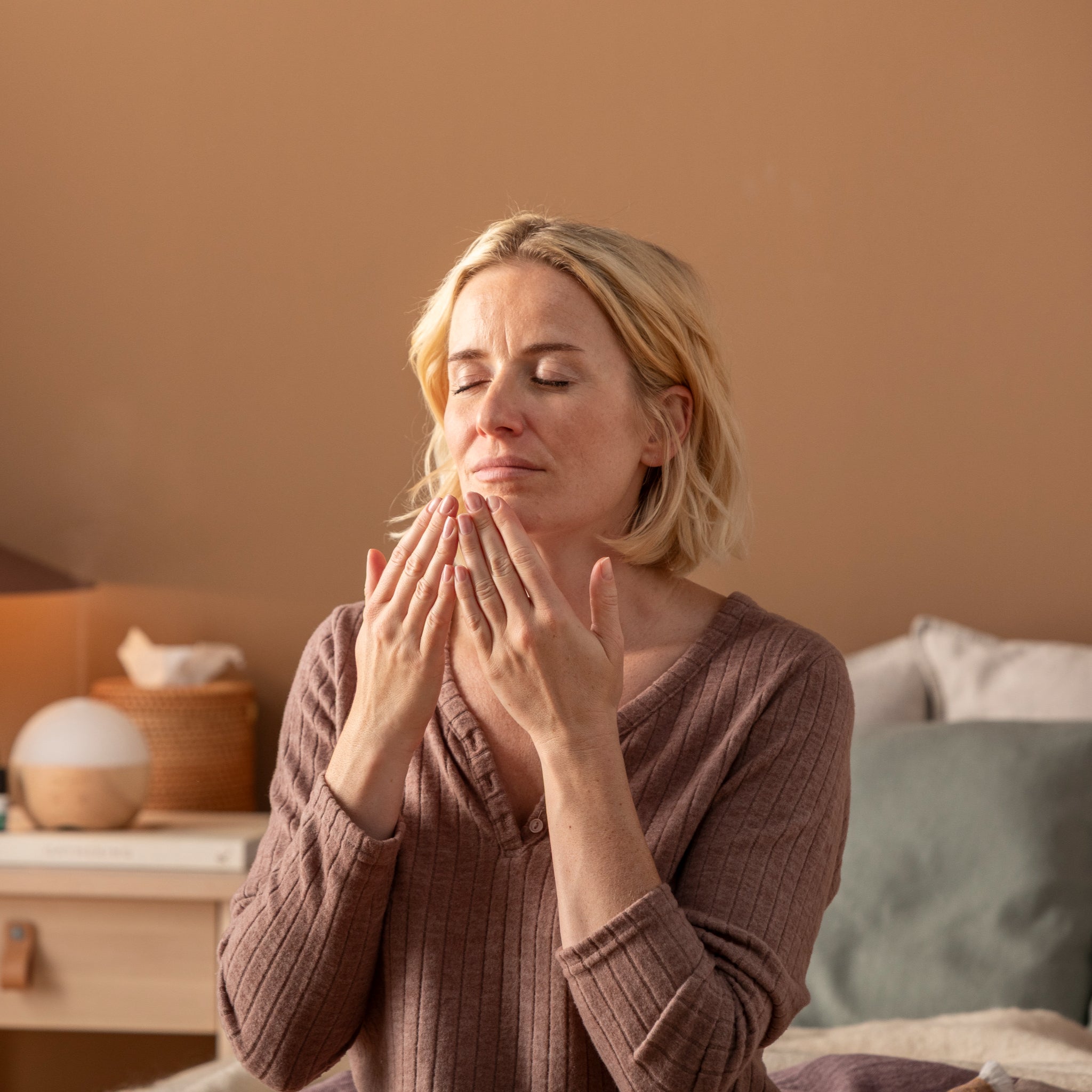 Eine Frau mit kurzen blonden Haaren sitzt mit geschlossenen Augen in einem Raum, hält die Hände vor das Gesicht und scheint einen beruhigenden Duft zu genießen. Der Raum hat warme Beigetöne und eine gemütliche, entspannte Atmosphäre.
