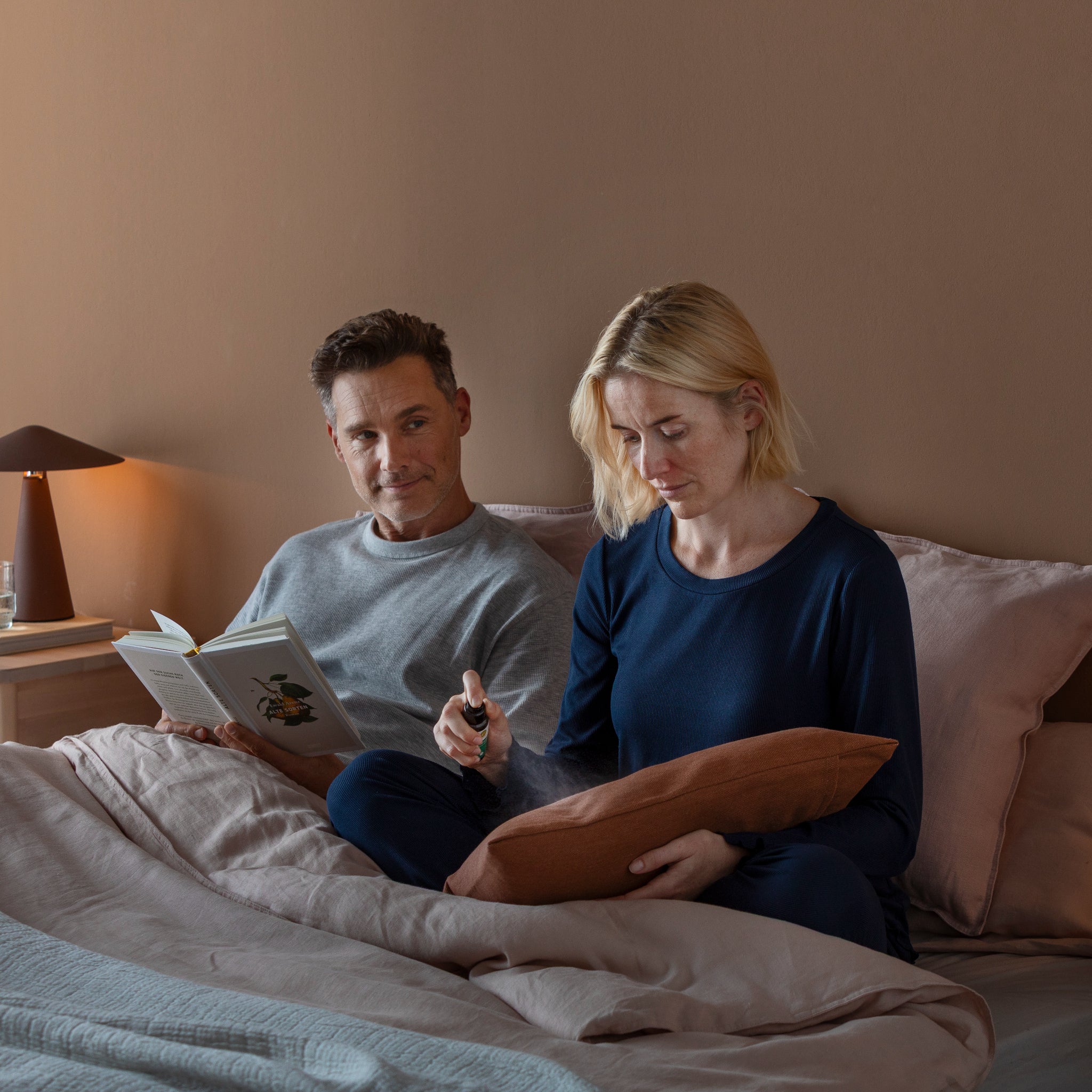 Ein Mann und eine Frau sitzen in einem Bett mit hellem Bettzeug. Der Mann im grauen Hemd liest in einem Buch und sieht die Frau an. Die Frau, in einem marineblauen Oberteil, sprüht etwas auf ein braunes Kissen auf ihrem Schoß. Eine Lampe und ein Glas Wasser stehen auf dem Nachttisch.
