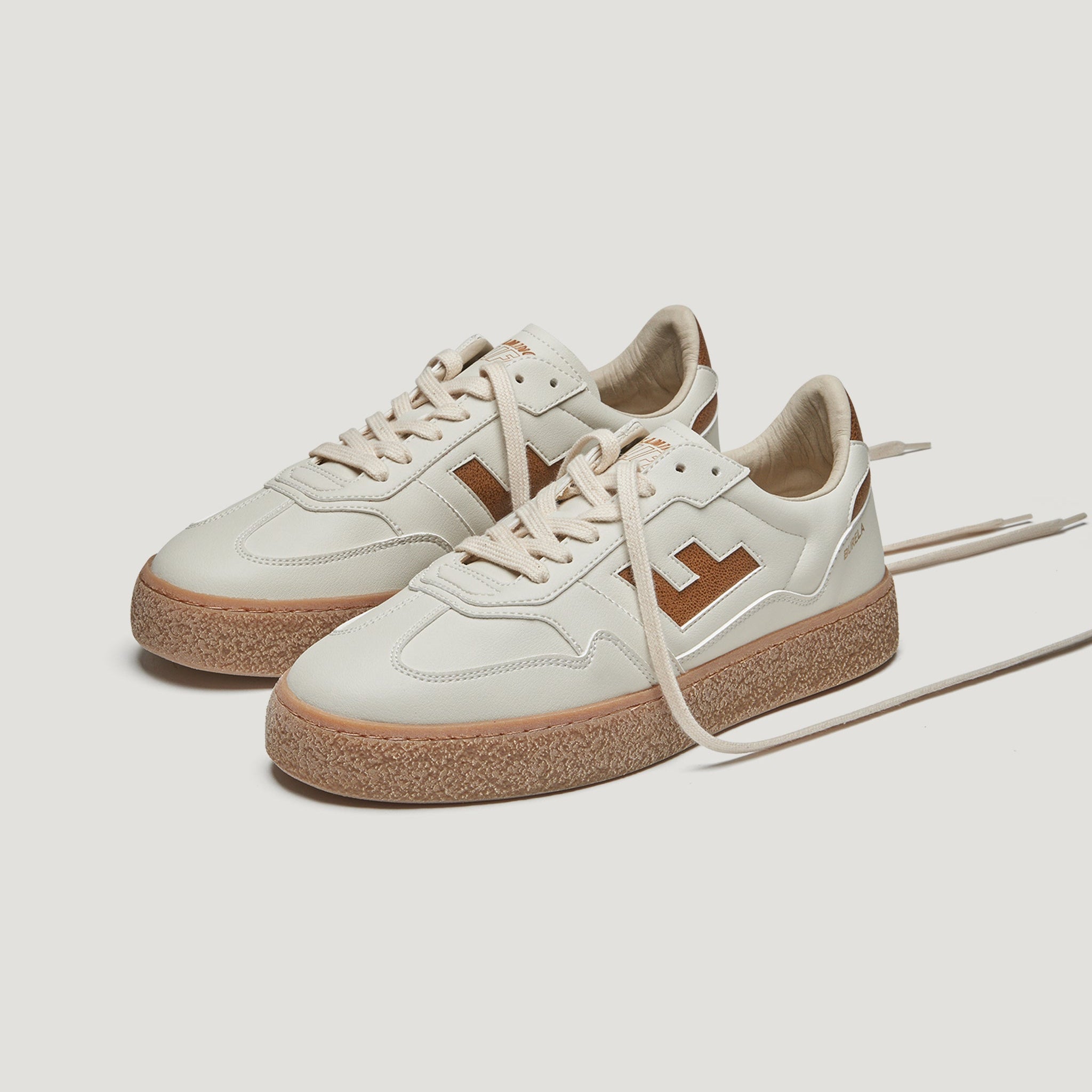 Ein Paar cremefarbene Turnschuhe mit braunen Gummisohlen, hellbeigen Schnürsenkeln und braunen geometrischen Logos an den Seiten. Die Schuhe sind auf einem weißen Hintergrund platziert, wobei die Schnürsenkel locker ausgelegt sind.