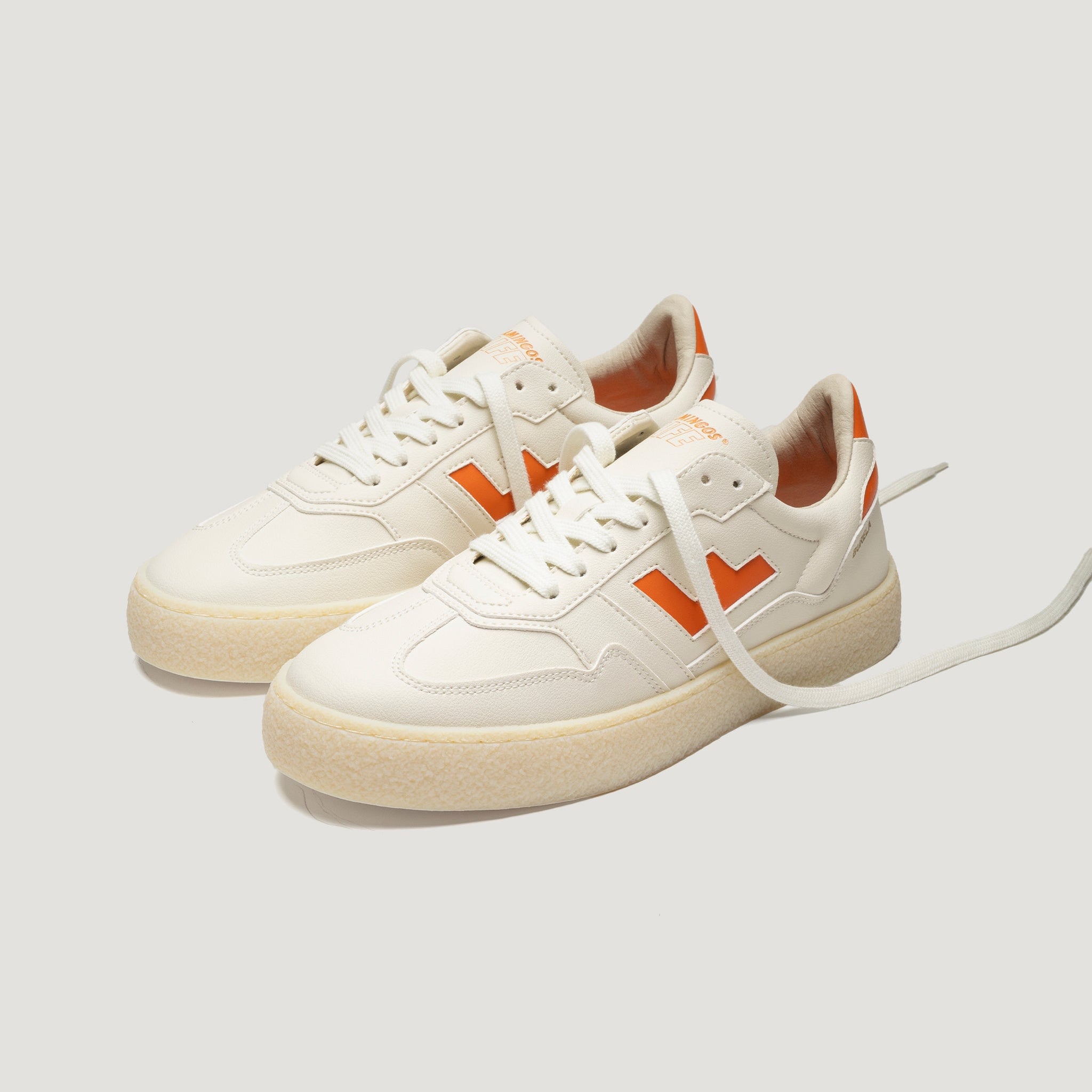 Ein Paar weißer Turnschuhe mit orangefarbenen Akzenten, orangefarbenen V-Logos an den Seiten, beigen Struktursohlen und weißen Schnürsenkeln, dargestellt auf einem schlichten, hellen Hintergrund.