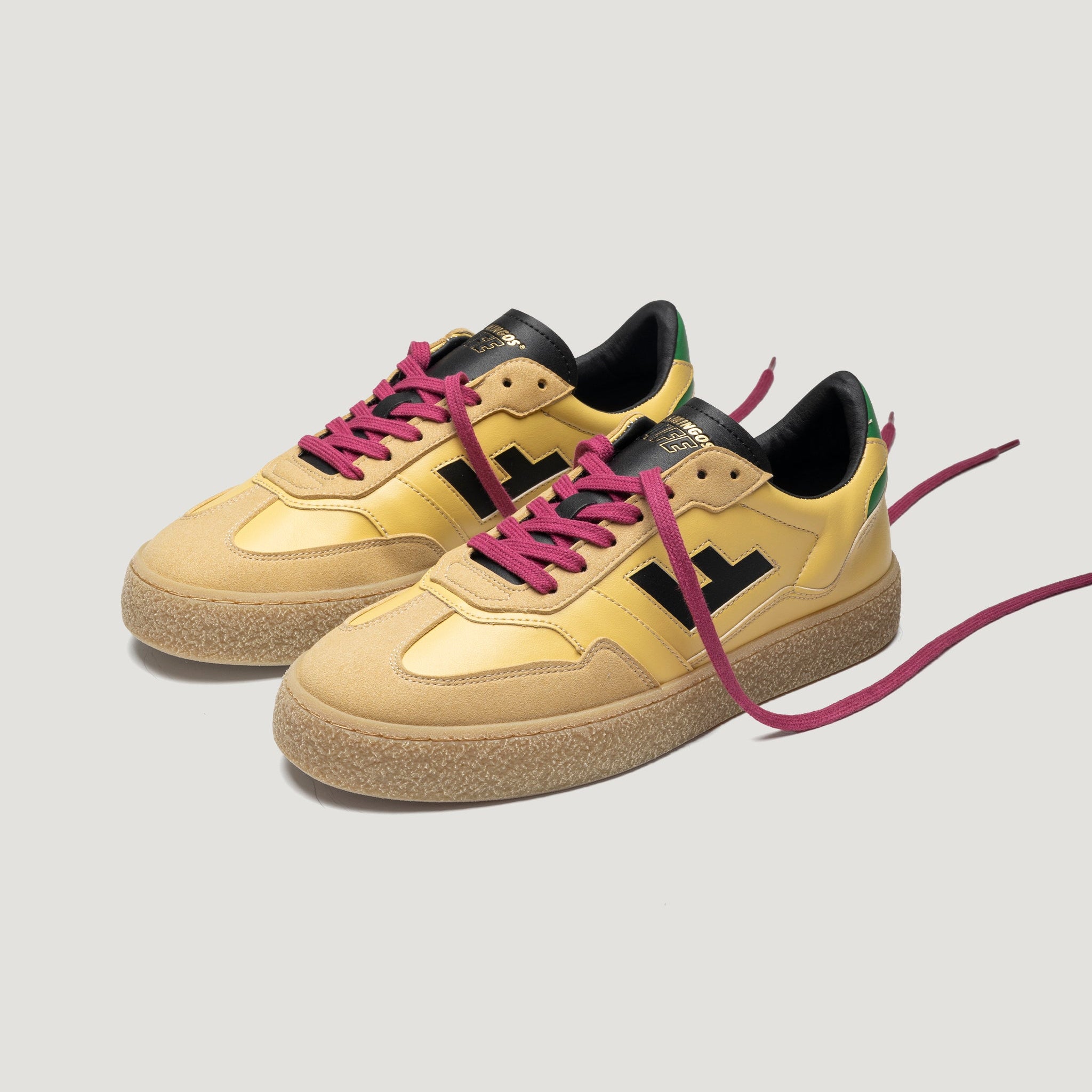 Ein Paar gelbe Turnschuhe mit hellbraunen Sohlen, schwarzen Akzenten und fetten schwarzen "H"-Logos an den Seiten. Sie haben dunkelrosa Schnürsenkel und schwarze Zungen, mit einem grünen Detail auf der Rückseite Ferse Bereich.