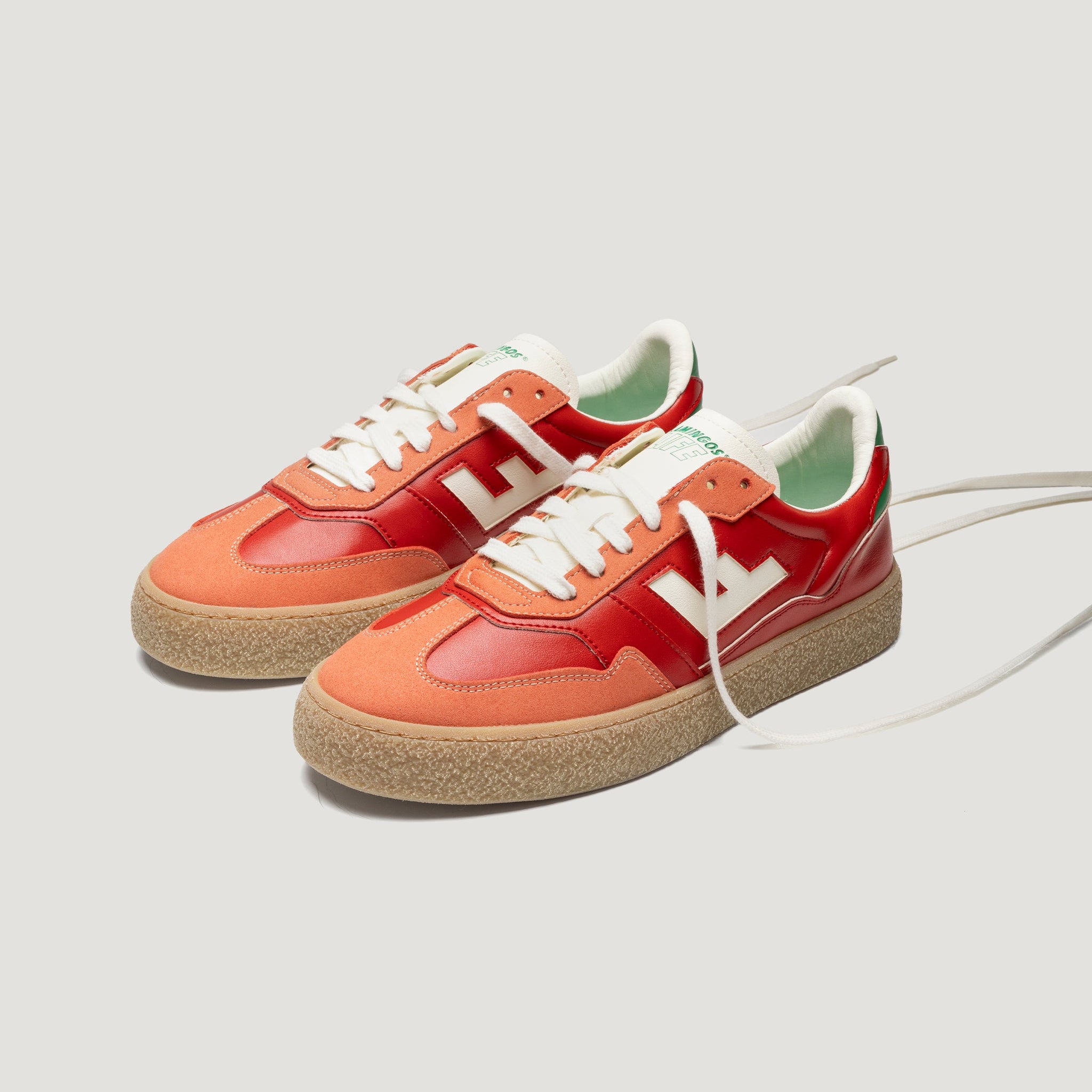 Ein Paar modische Turnschuhe mit orangefarbenen und roten Einsätzen, weißen Schnürsenkeln, beigen Sohlen und einem weißen L-Motiv an den Seiten auf einem hellgrauen Hintergrund.