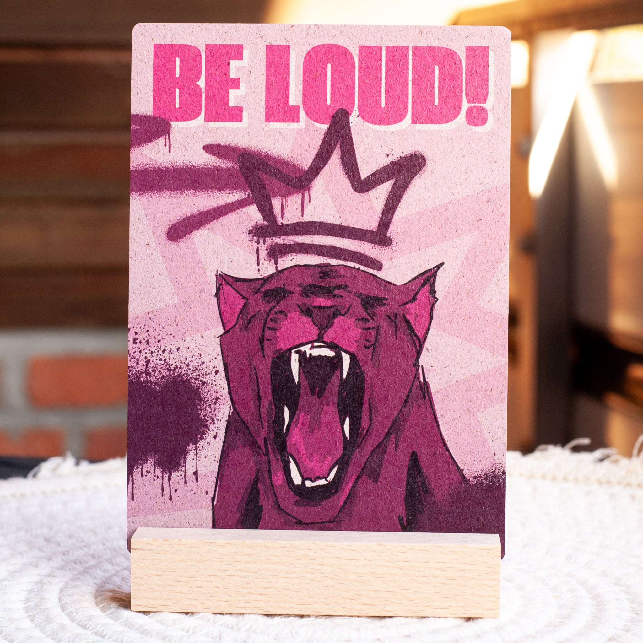 Ein Schild mit der fettgedruckten rosafarbenen Aufschrift BE LOUD! über einem illustrierten brüllenden Panther und einer aufgesprühten Krone auf rosafarbenem Hintergrund, aufgestellt auf einem Holzständer.