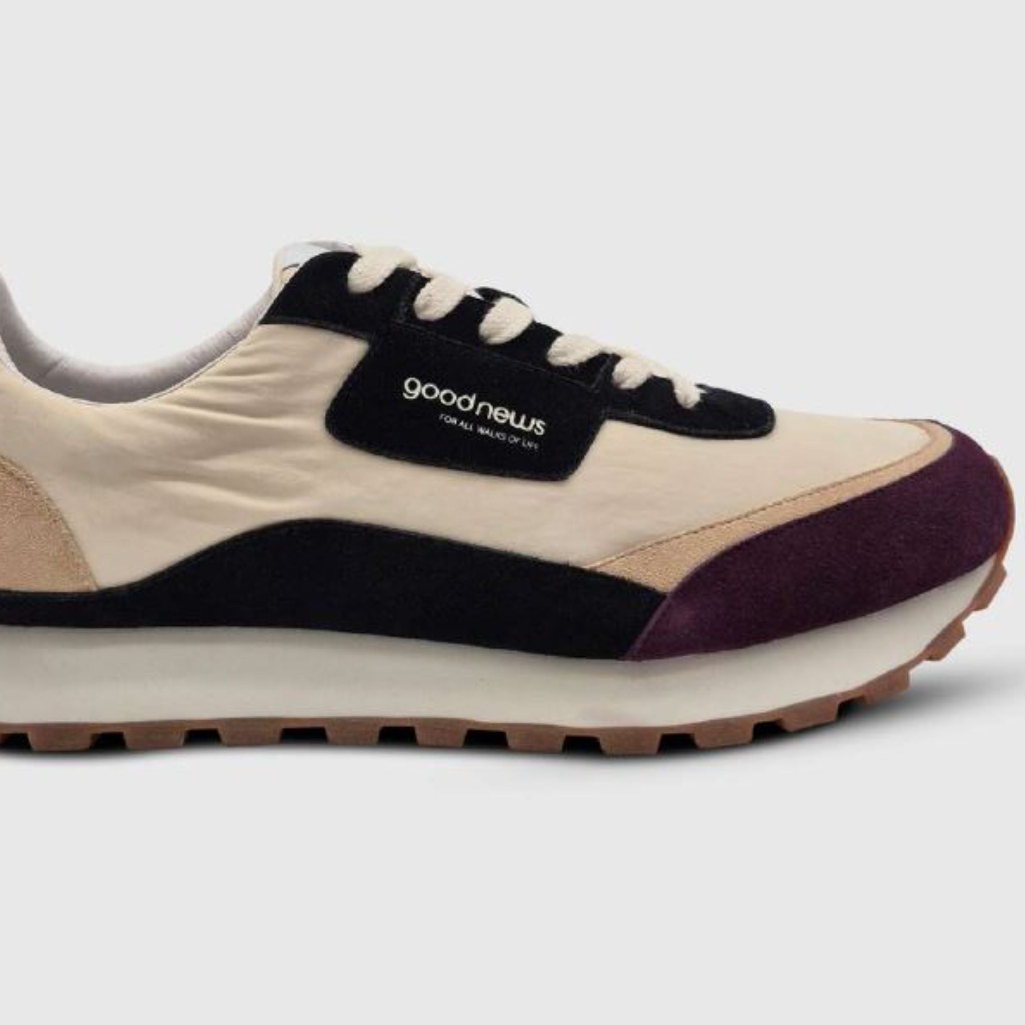 Ein beiger, schwarzer und lila Sneaker mit Gummisohle und schwarzen Schnürsenkeln. An der Seite befindet sich ein Good-News-Logo-Tag vor einem schlichten, hellen Hintergrund.