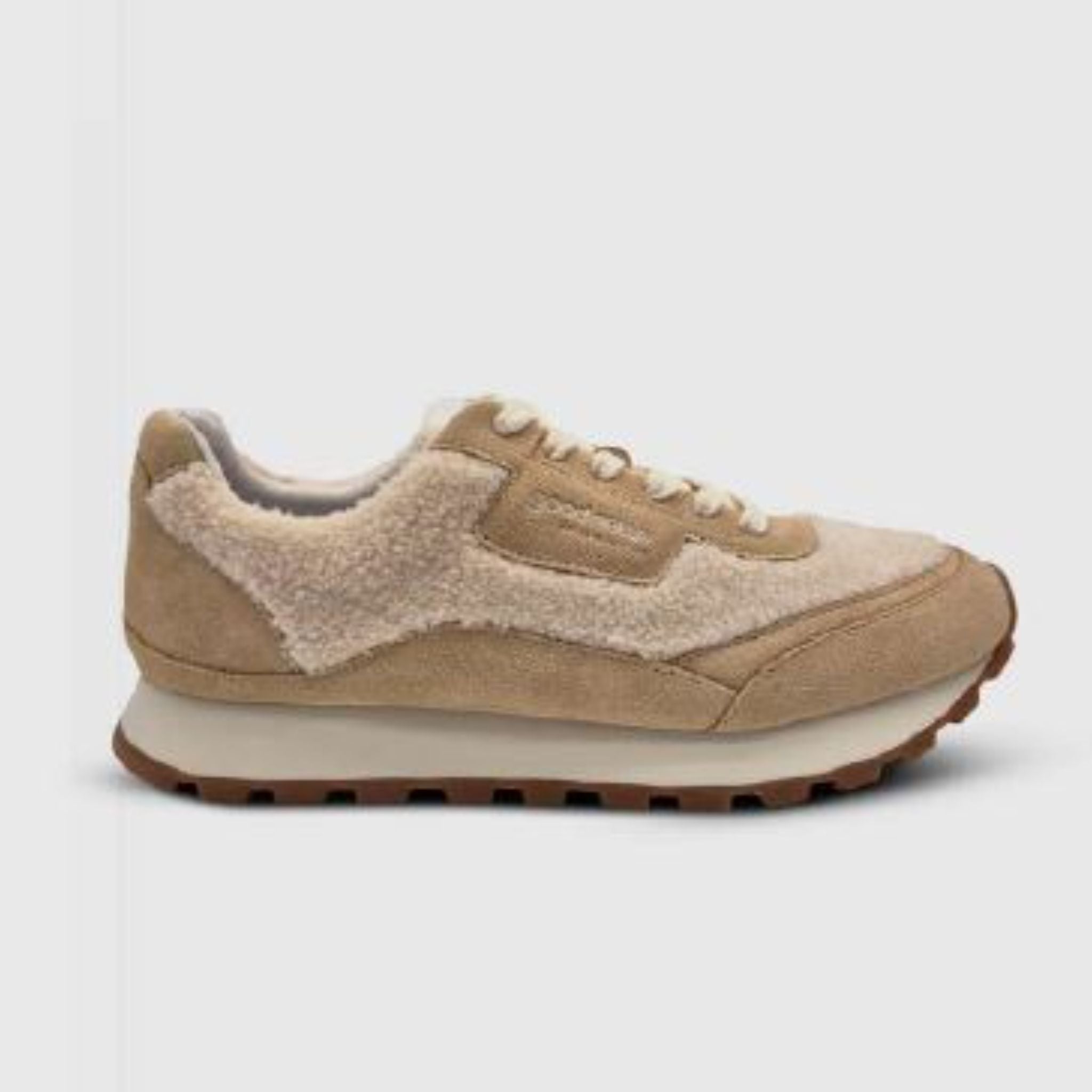 Ein beiger Sneaker mit Wildleder- und Shearling-Texturen, einer dicken Gummisohle mit braunem Profil, weißen Schnürsenkeln und einem tief ausgeschnittenen Knöchel, der auf einem einfarbigen grauen Hintergrund steht.