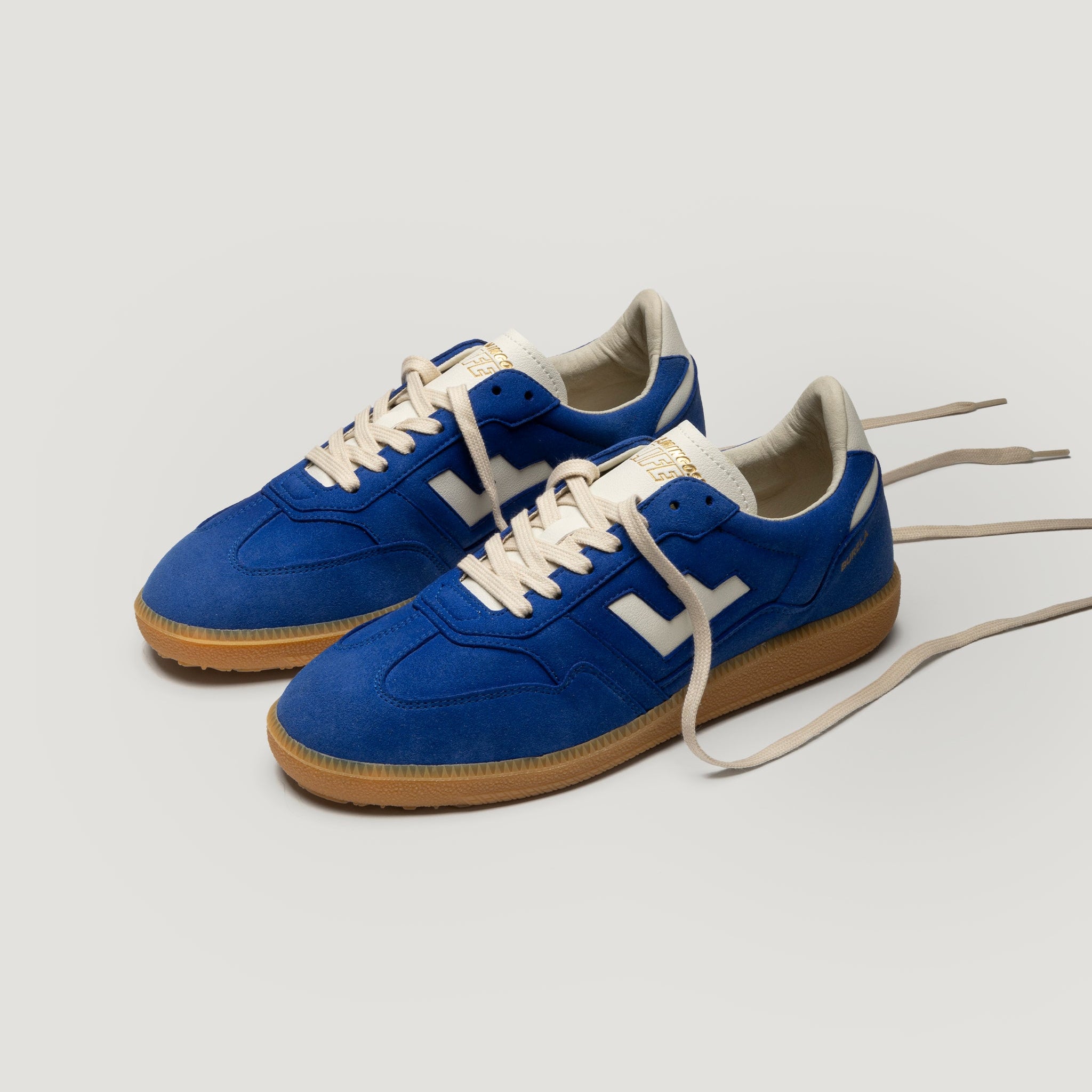 Ein Paar blaue Wildleder-Sneakers mit beigen Schnürsenkeln, weißen Details und Gummisohlen auf einem hellgrauen Hintergrund.