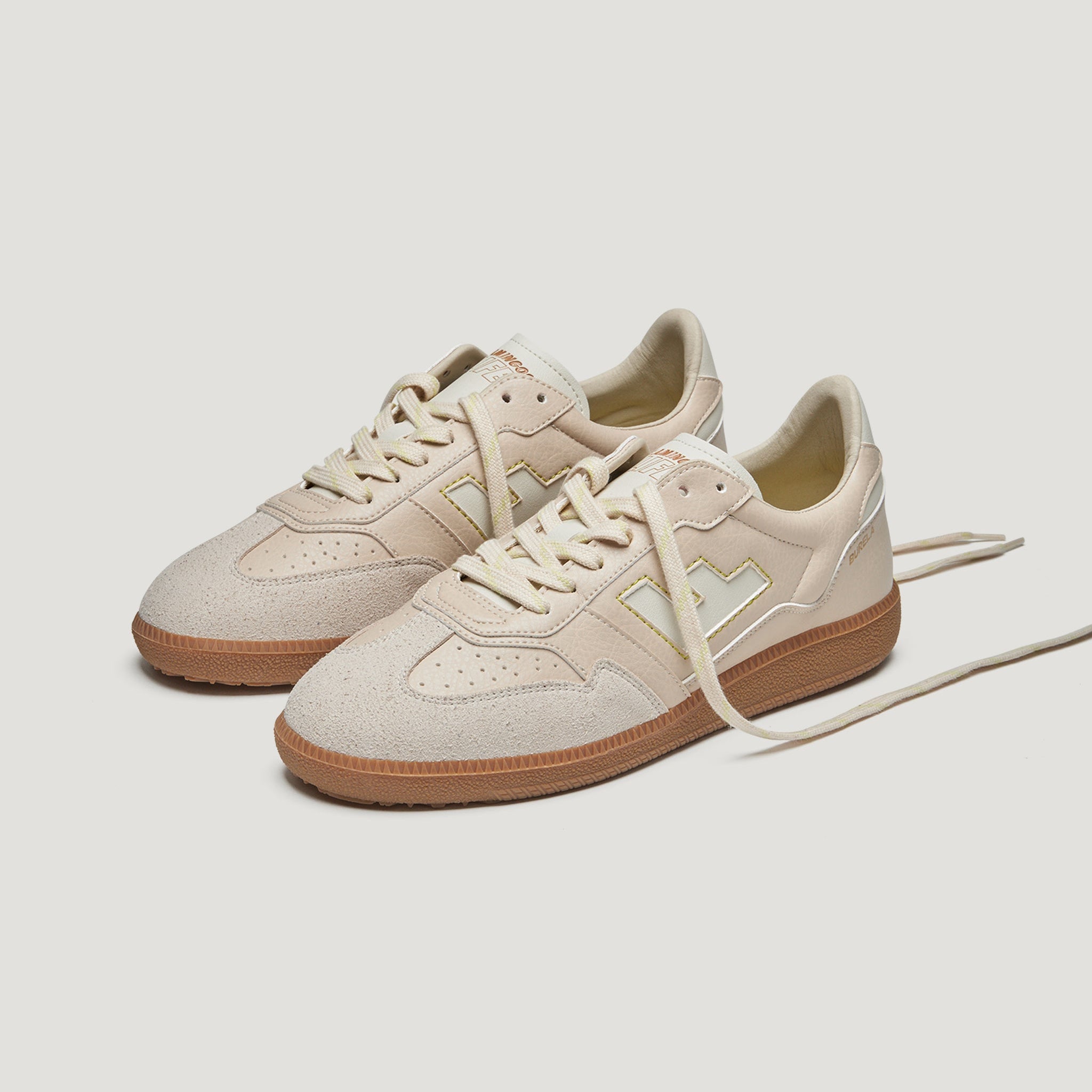 Ein Paar beigefarbene Turnschuhe mit weißen Schnürsenkeln, beigen Seitenteilen mit geometrischem Muster und hellbraunen Gummisohlen auf einem einfarbig weißen Hintergrund.