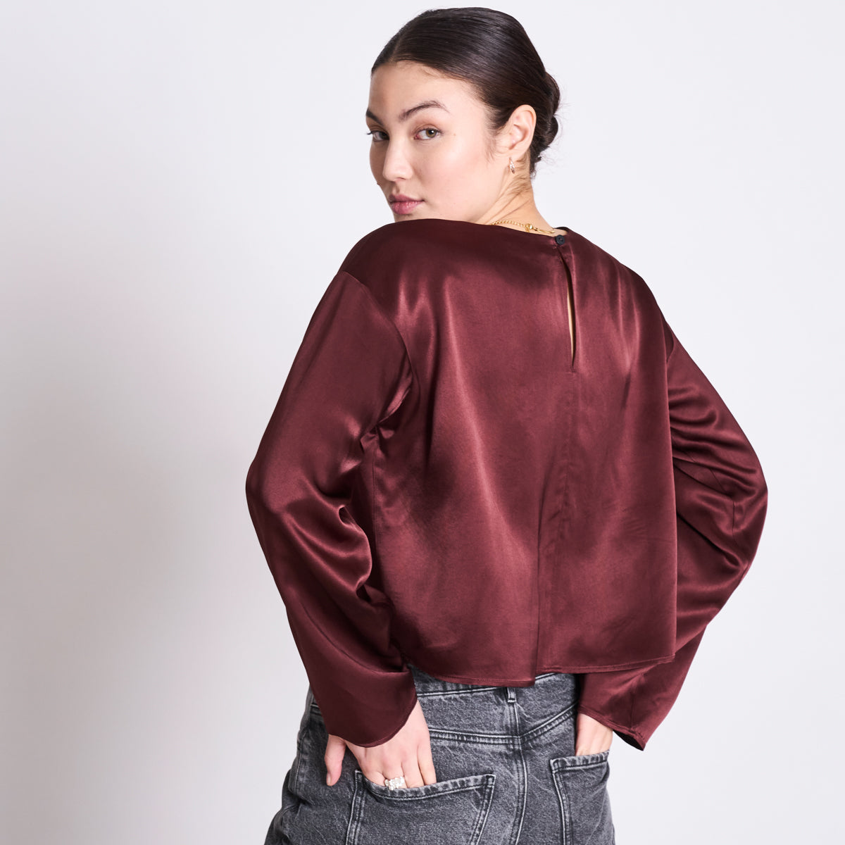 Eine Frau mit dunklen Haaren in einem tiefen Dutt, die eine glänzende kastanienbraune langärmelige Bluse und graue Jeans mit hoher Taille trägt, steht mit dem Rücken zur Kamera und schaut über ihre Schulter.